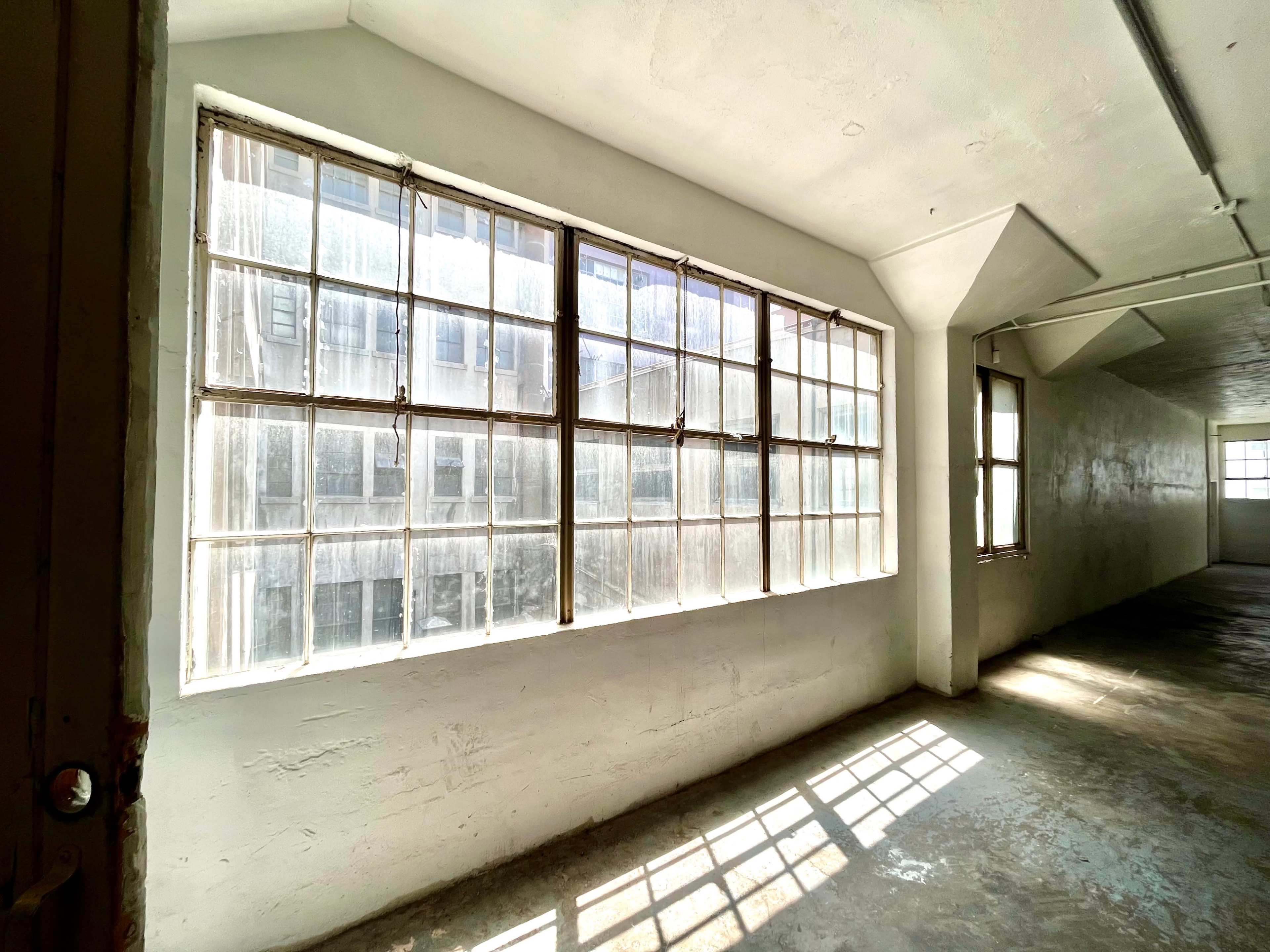 A row of large, dusty windows lines one wall of a vacant, sunlit room with concrete floors.