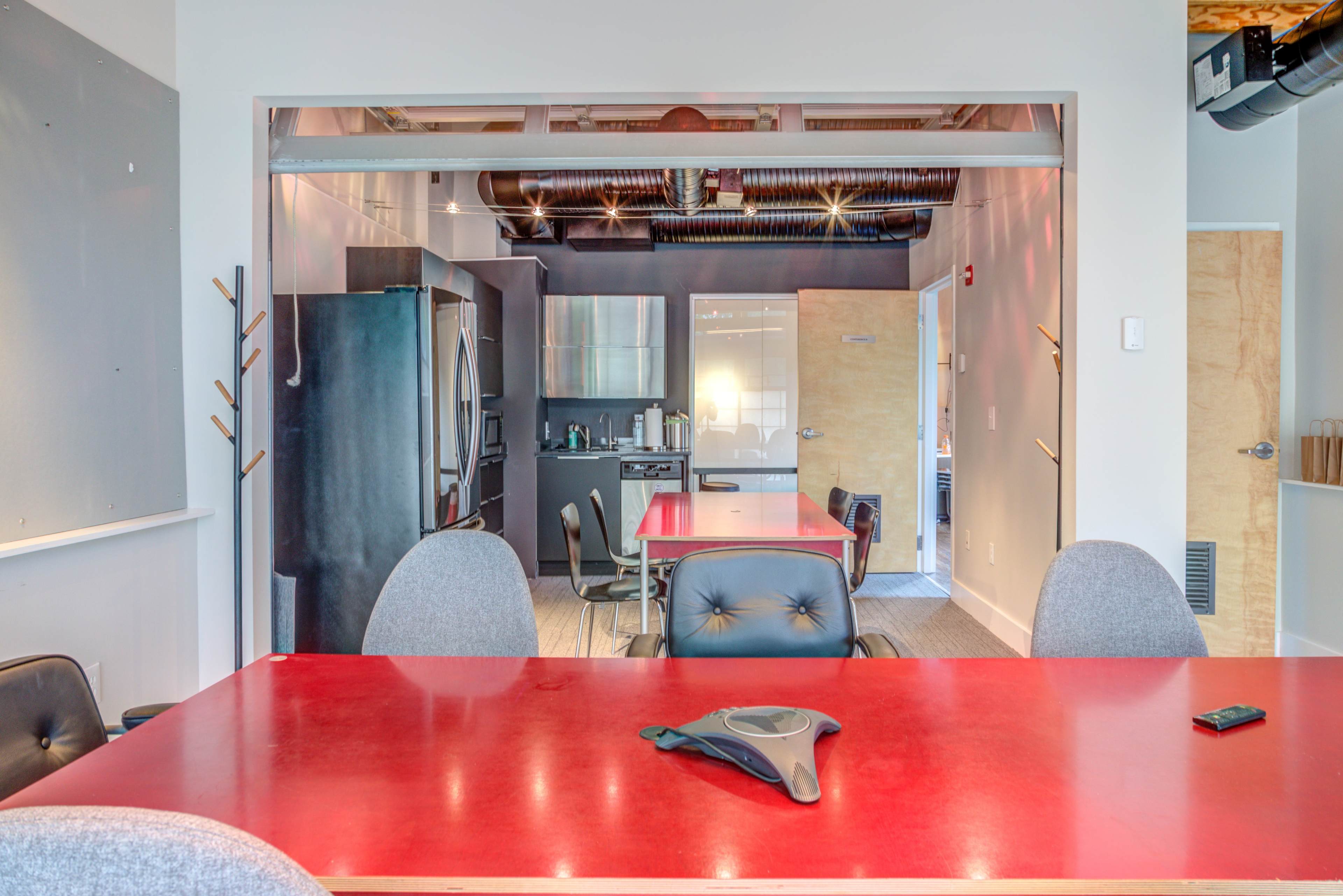 A red conference table is set in the foreground, with gray chairs arranged around it, leading to a kitchen area visible in the background.