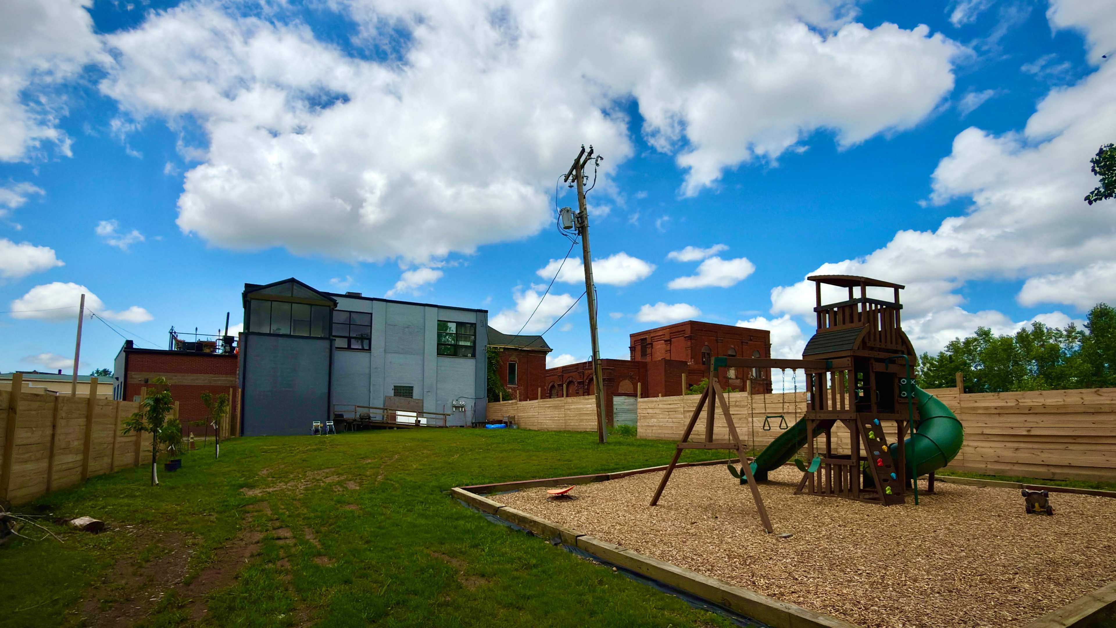 The image shows a playground with a wooden swing set and slide in a grassy area, surrounded by wooden fencing and buildings under a blue sky with clouds.