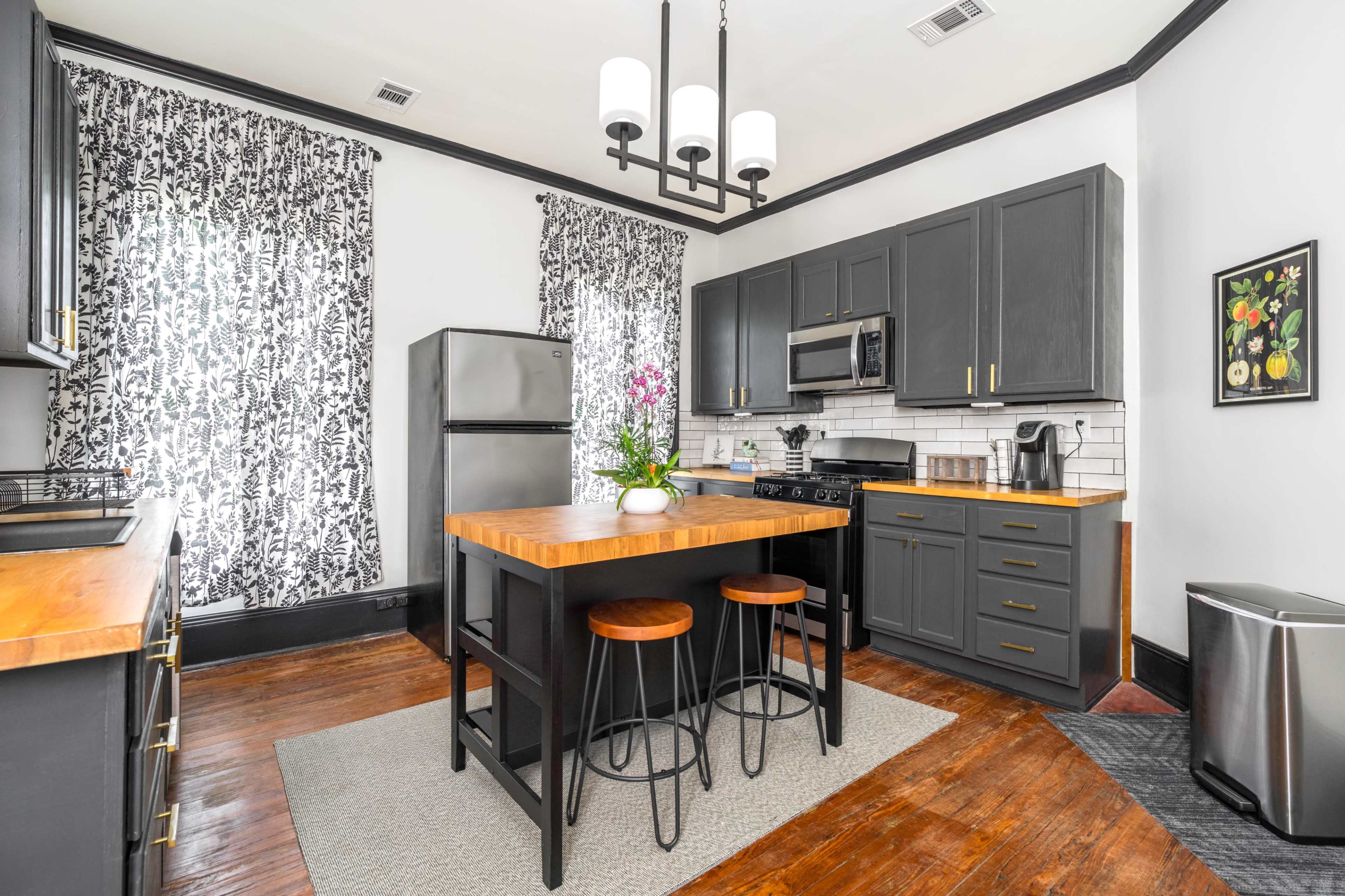 A modern kitchen features dark cabinetry, a central wooden island with two stools, and large windows adorned with patterned curtains.