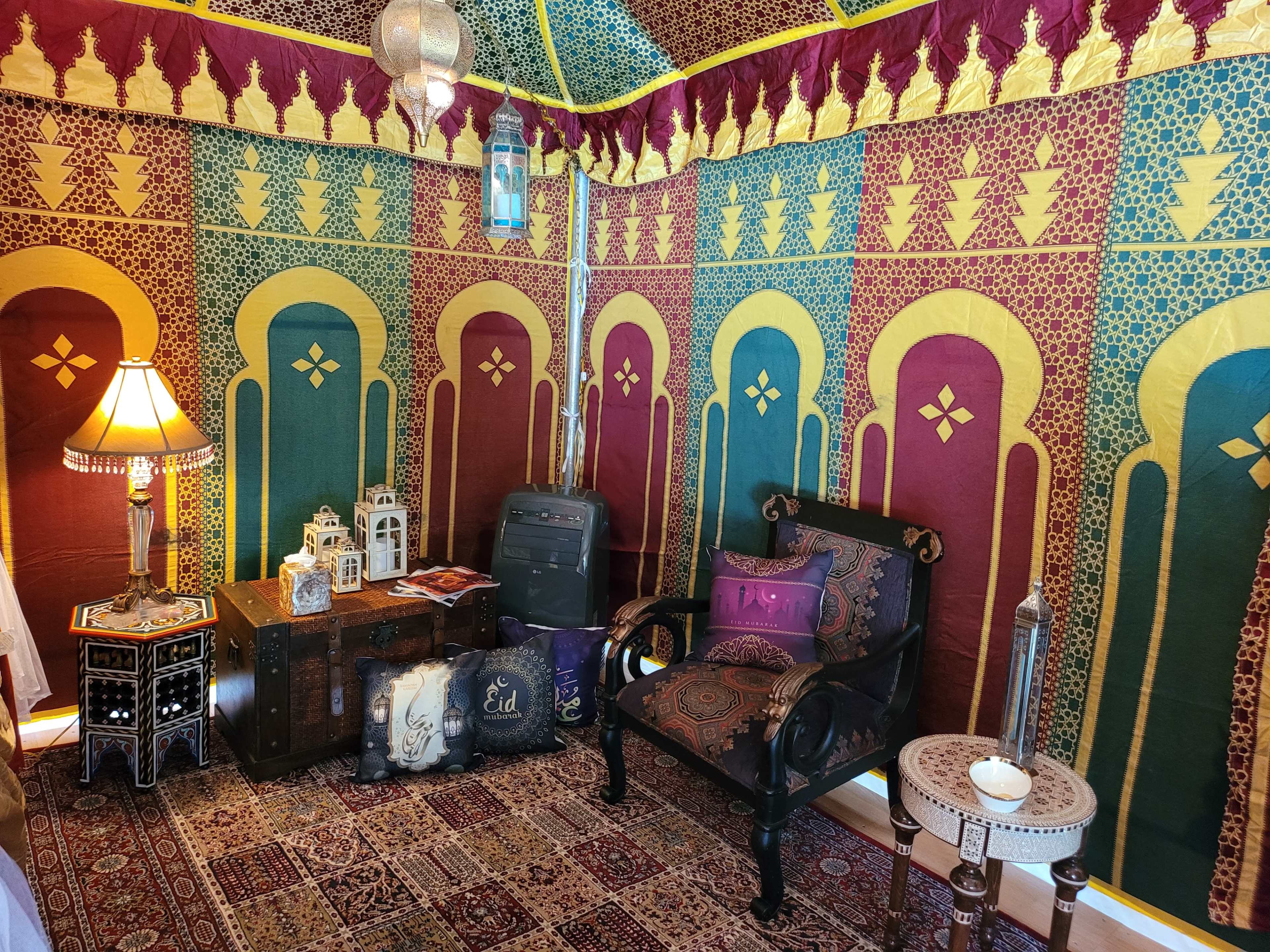 A colorful tent-like interior adorned with patterned fabrics, featuring a decorative seating area with a lamp and various ornamental items.