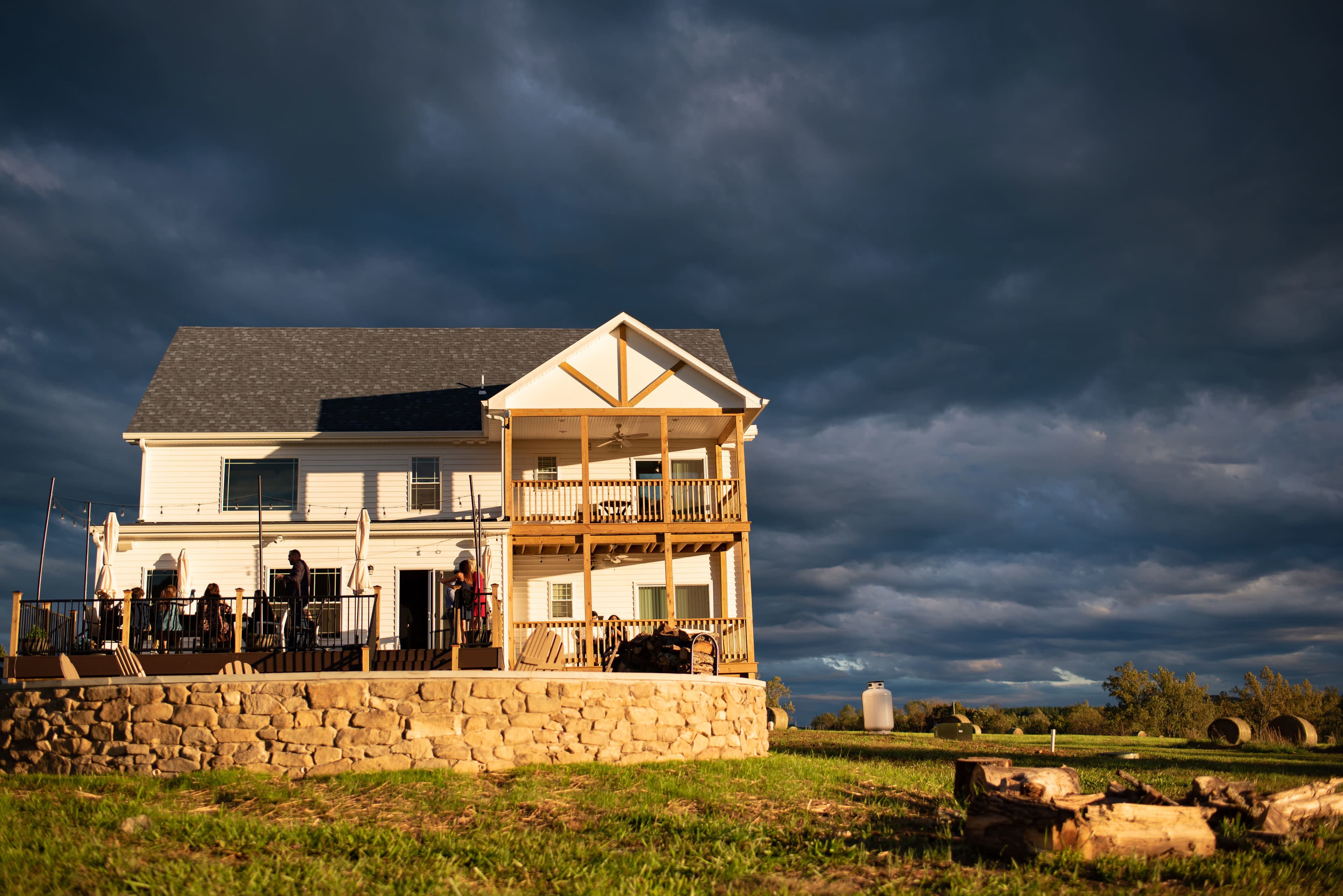 A two-story house with multiple balconies stands on a grassy hillside under a dramatic sky.