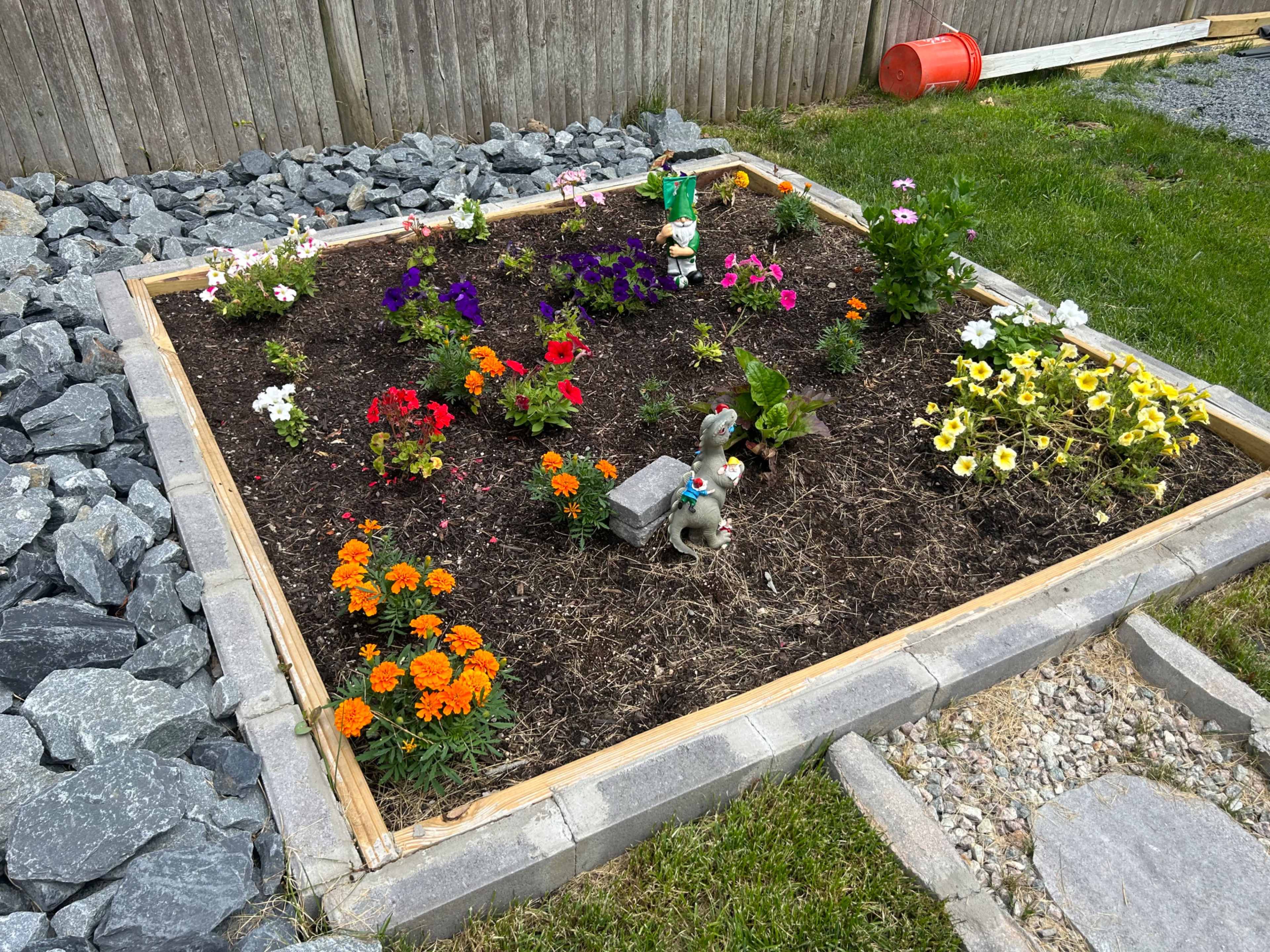 A flower garden is framed by wooden borders and contains various colorful flowers, a small statue, and decorative stones.