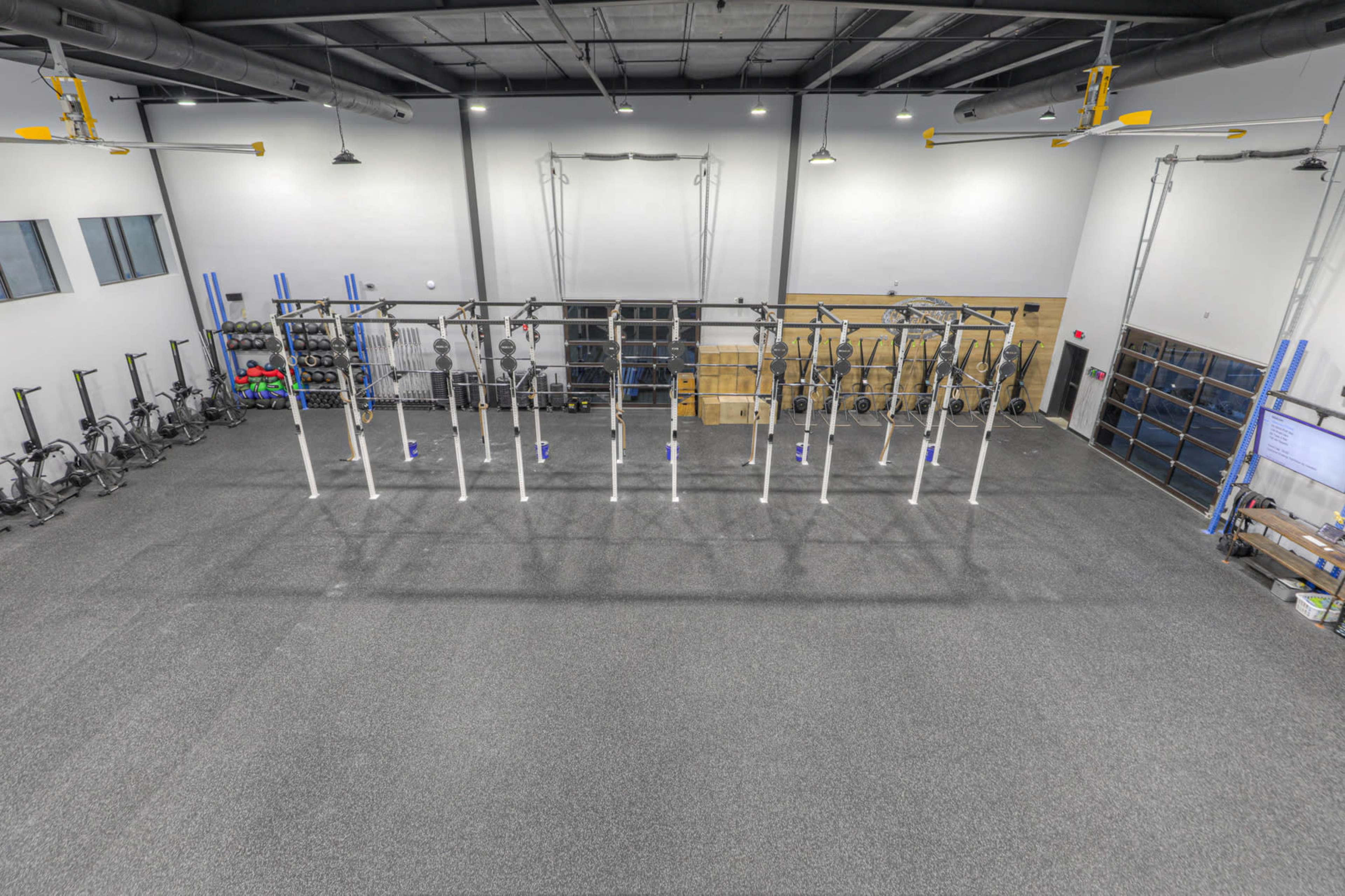 The image shows a large gym interior with various exercise equipment arranged around a central area featuring a pull-up rig and storage.