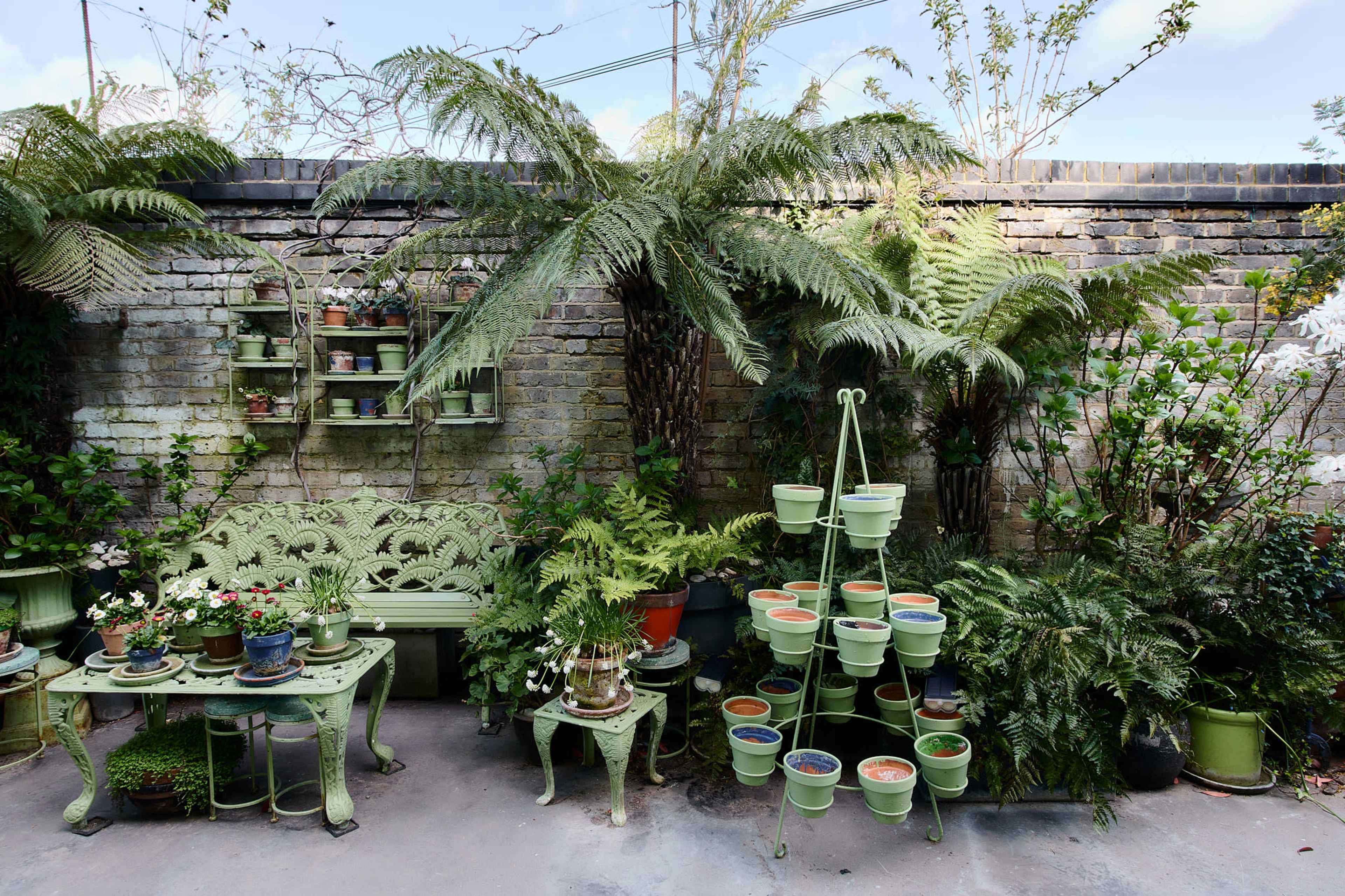 The image shows a lush garden area with a green bench and table surrounded by various plants and a tiered plant stand against a brick wall.