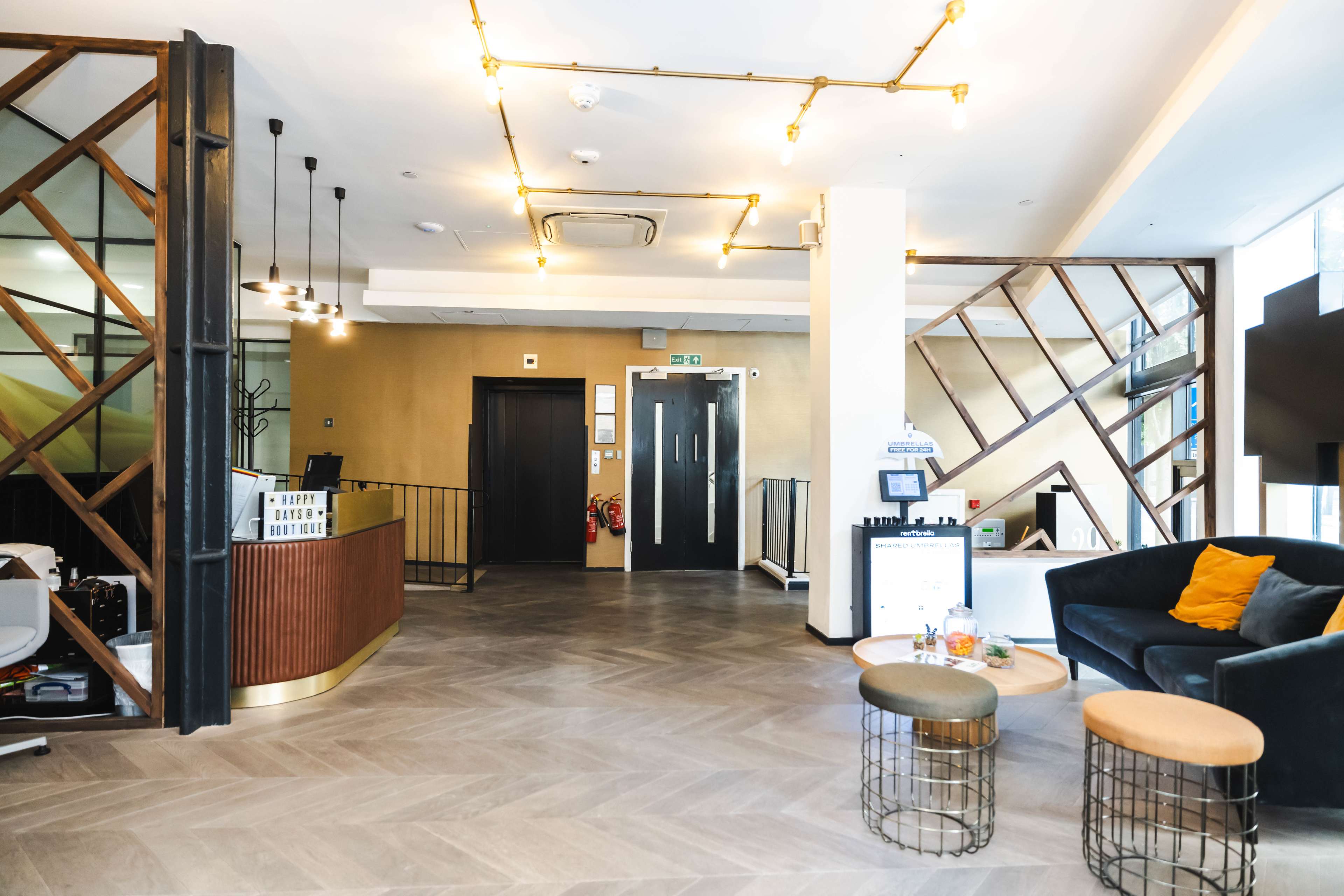 A modern hotel lobby featuring a reception desk, seating area with a black sofa, decorative lighting, and a staircase.