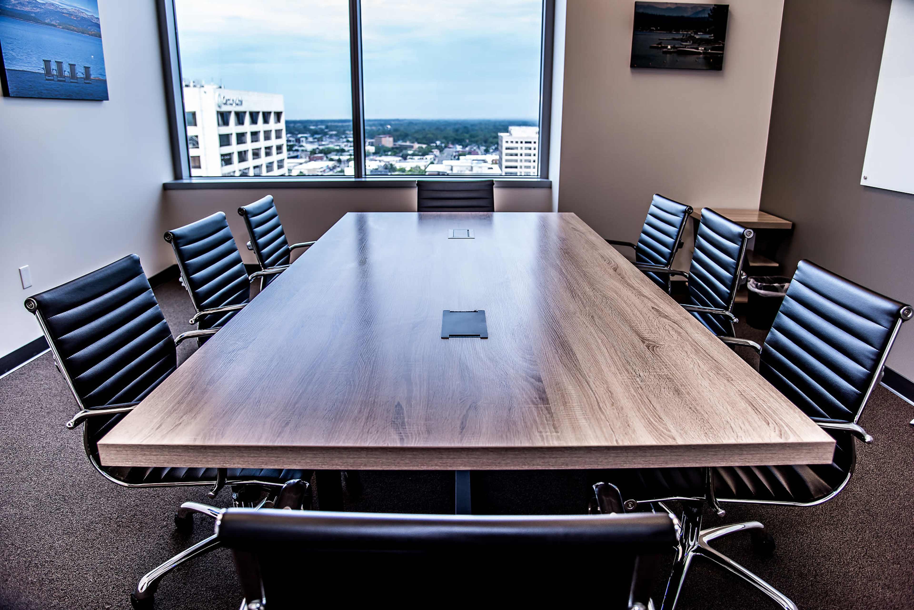 A rectangular conference table with a polished wooden surface is surrounded by black leather chairs in a high-rise office with large windows.