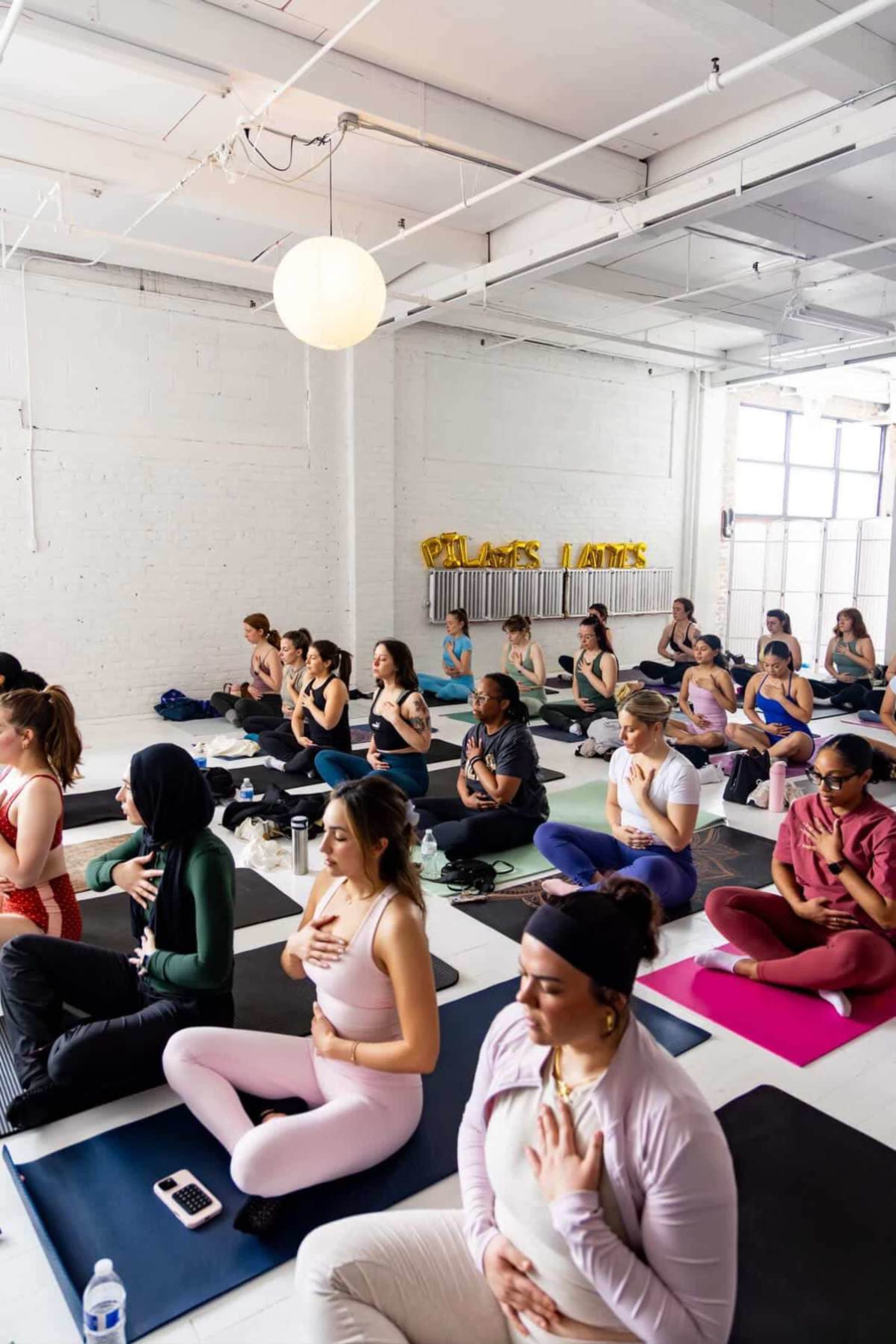 A group of individuals participates in a yoga class in a bright, spacious studio, seated on mats in a meditative pose.