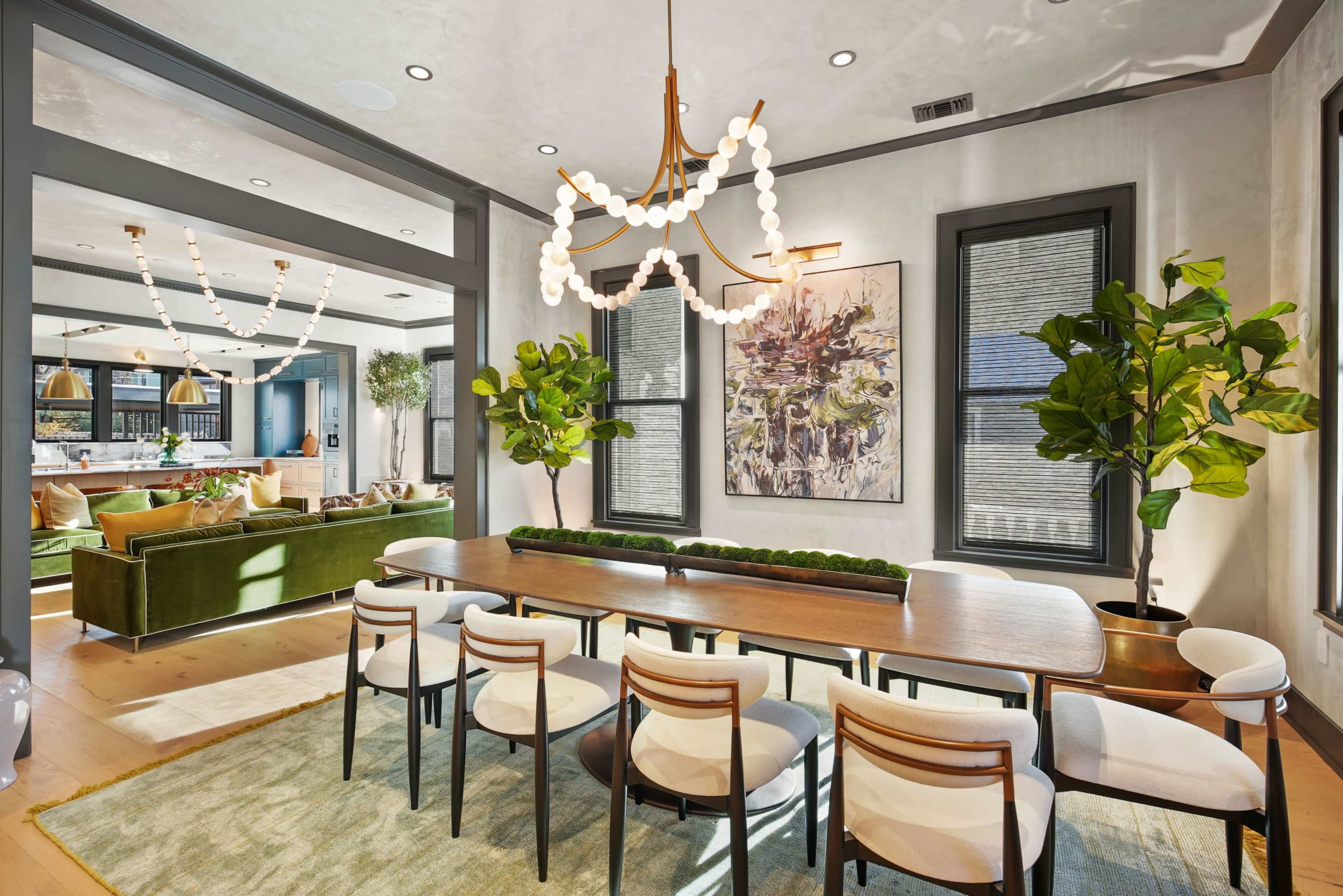 The image shows a brightly lit dining area featuring a long wooden table with eight chairs, surrounded by indoor plants and a decorative chandelier, with a visible living space in the background.