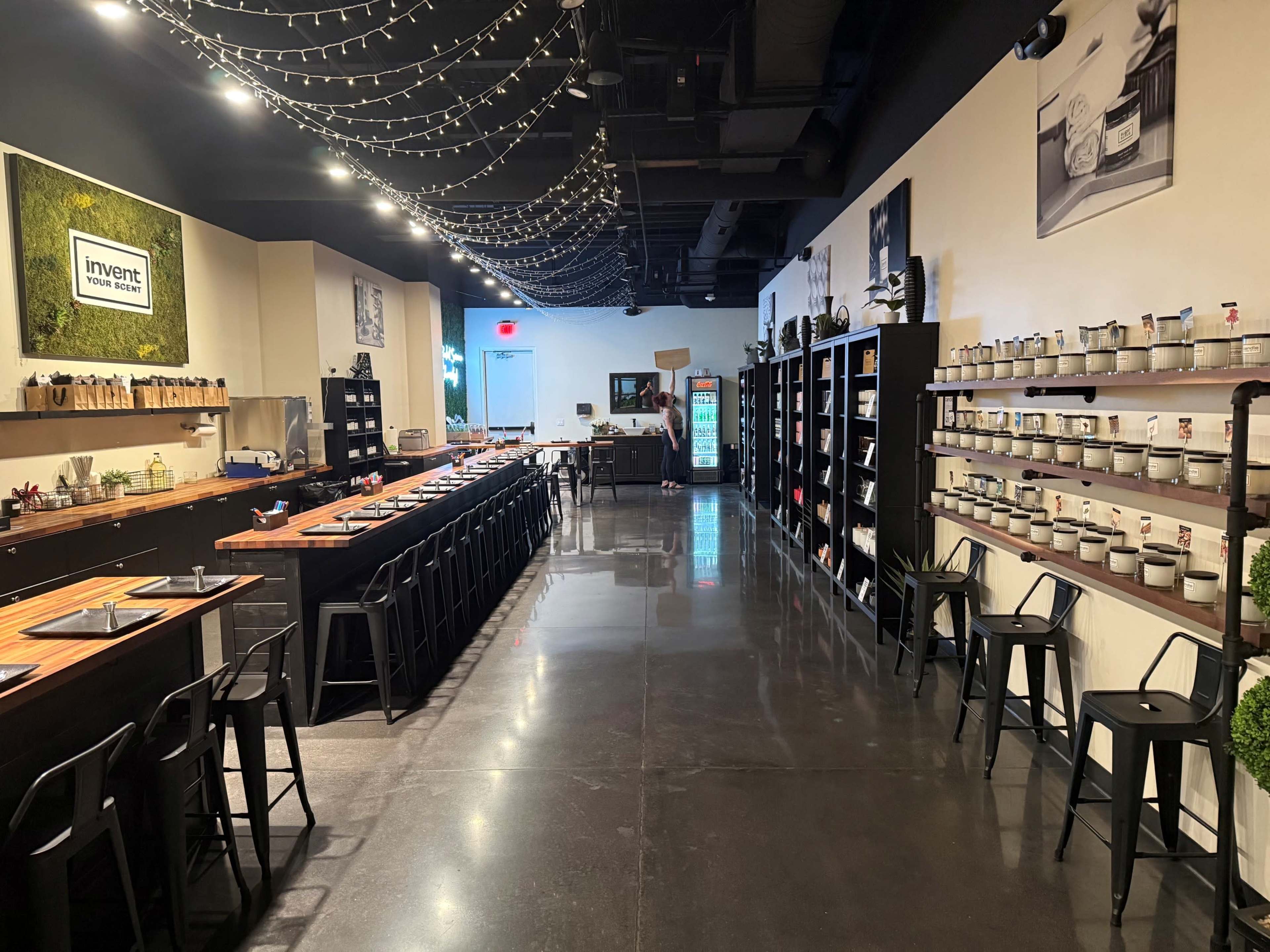 The image shows a modern kitchen space with wooden tables, black stools, and shelves lined with cooking supplies and ingredients under soft string lights.