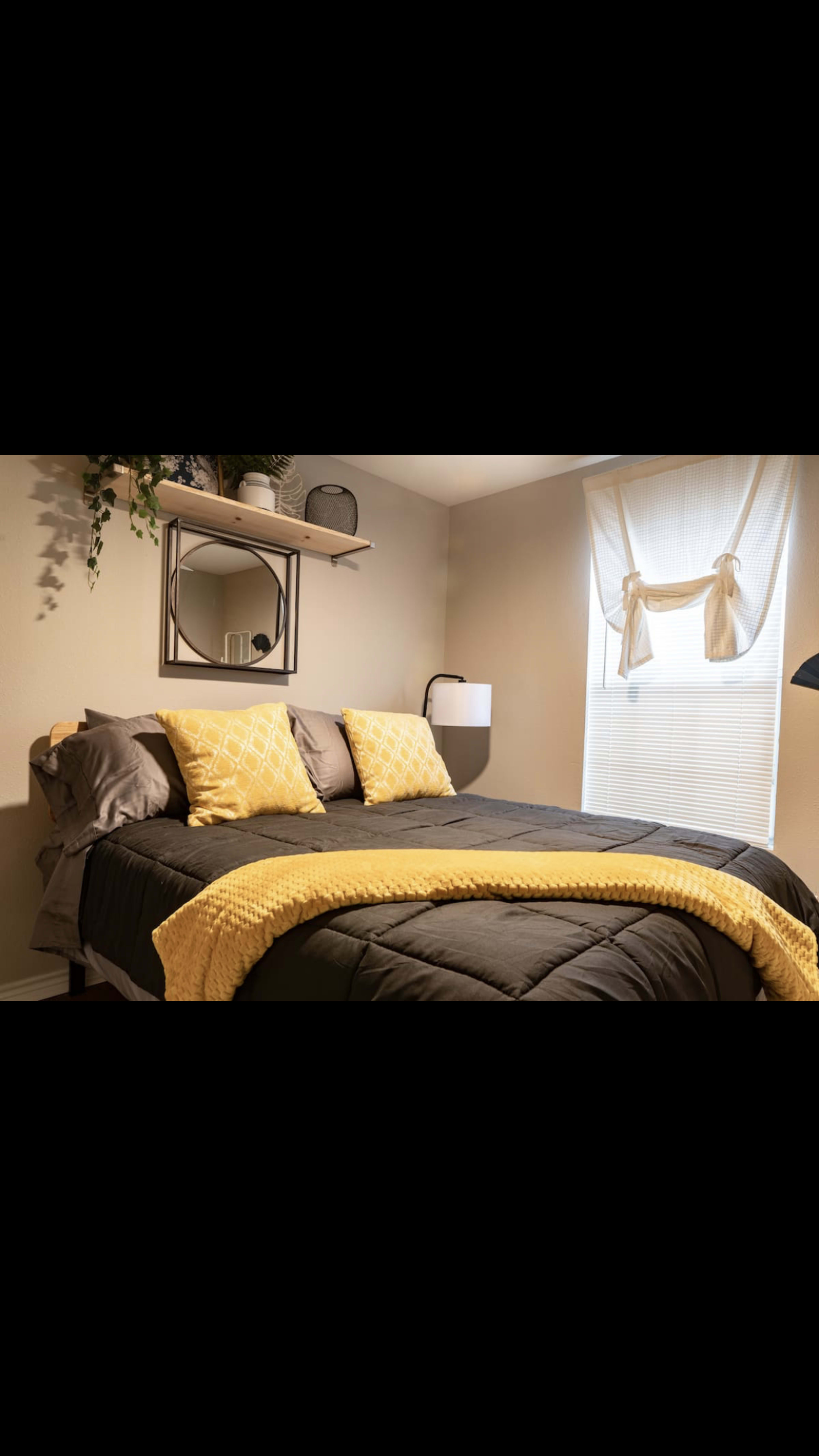 A neatly made bed with yellow and gray bedding, accented by a shelf above and natural light coming through a window with curtains.