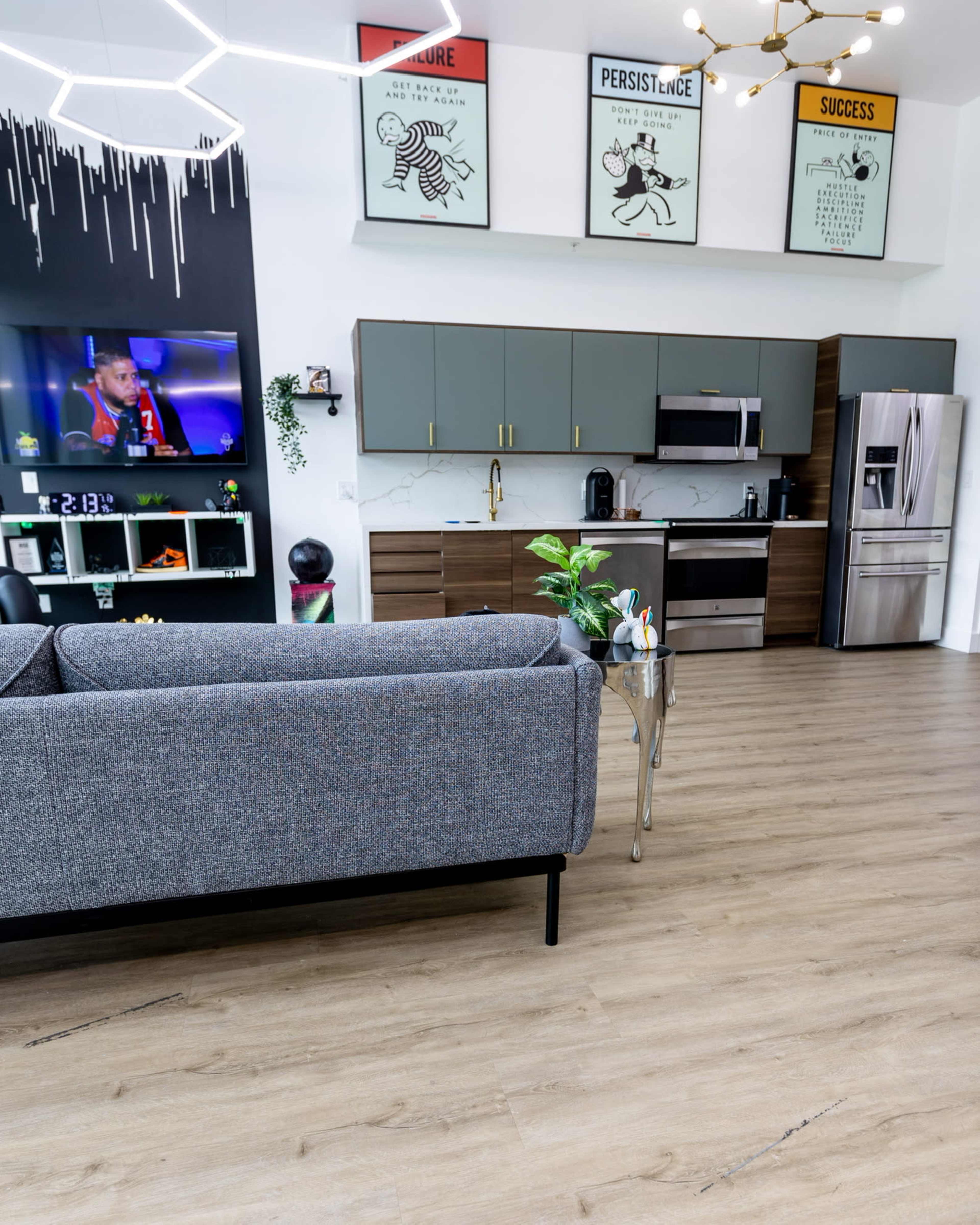 A modern kitchen and living area with green and brown cabinetry, a television displaying a sports event, and motivational artwork on the walls.