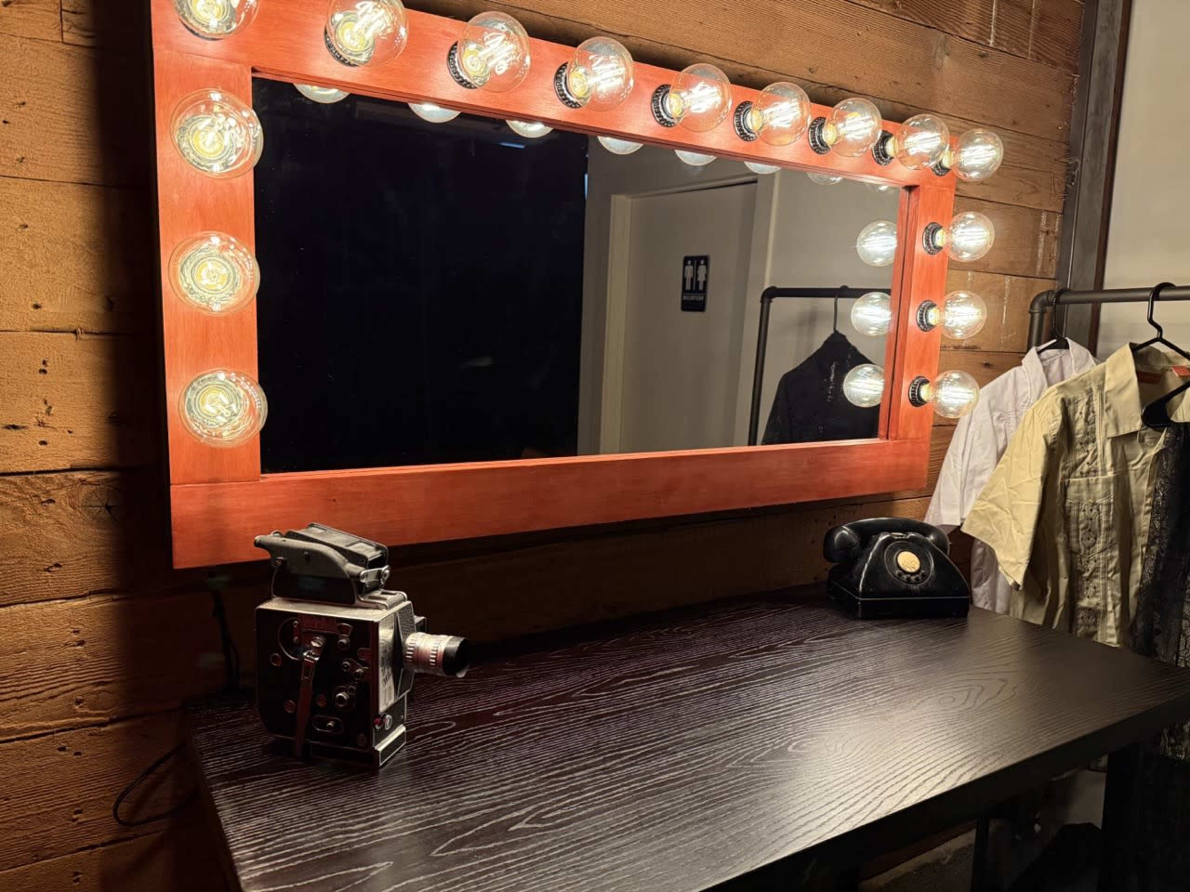 The image shows a wooden vanity with a large, illuminated mirror, a vintage camera, an old-fashioned telephone, and hanging clothes in the background.