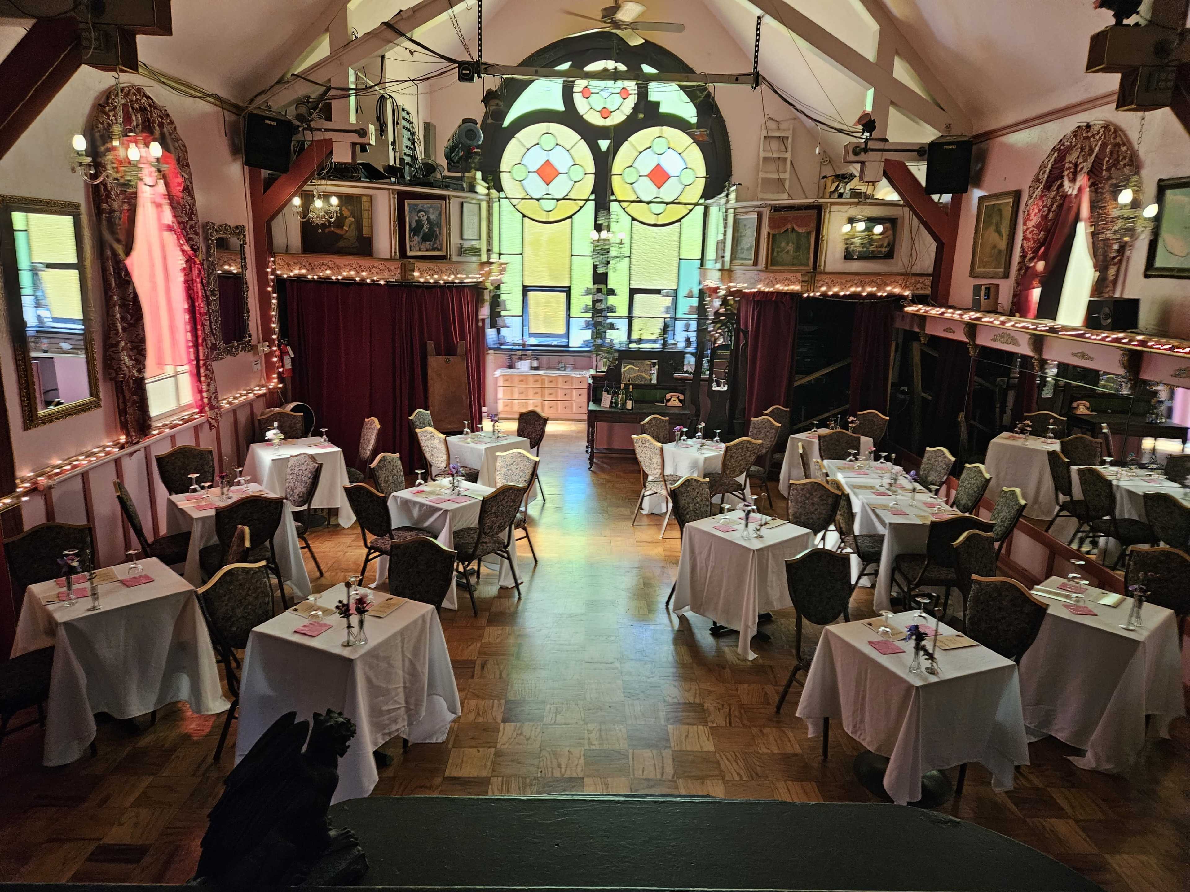 The image shows a spacious dining room with neatly set tables and a large stained glass window at the far end.
