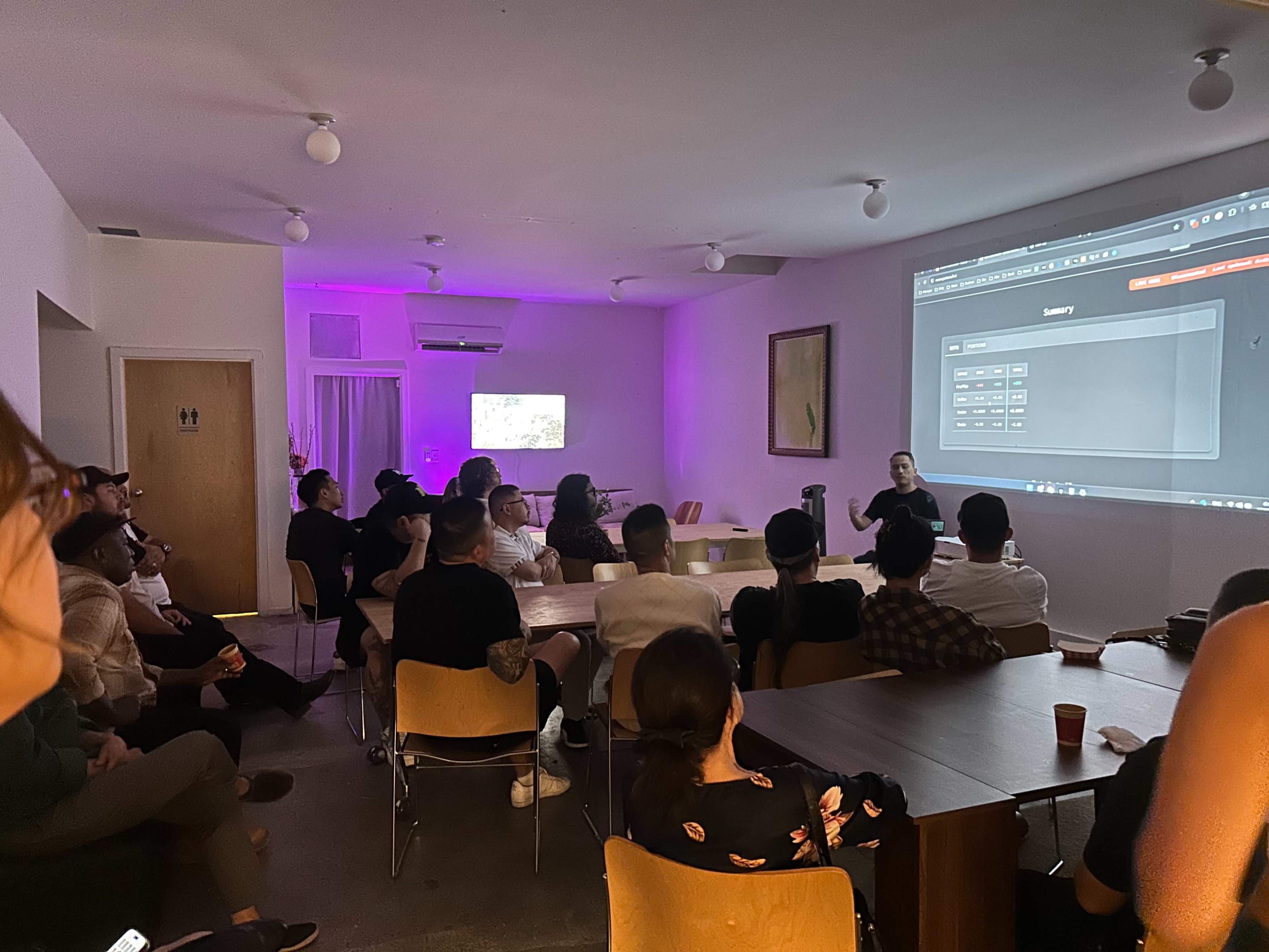 A group of people sits in a room with dim lighting, watching a presentation projected on a screen while a speaker stands in front, engaging the audience.