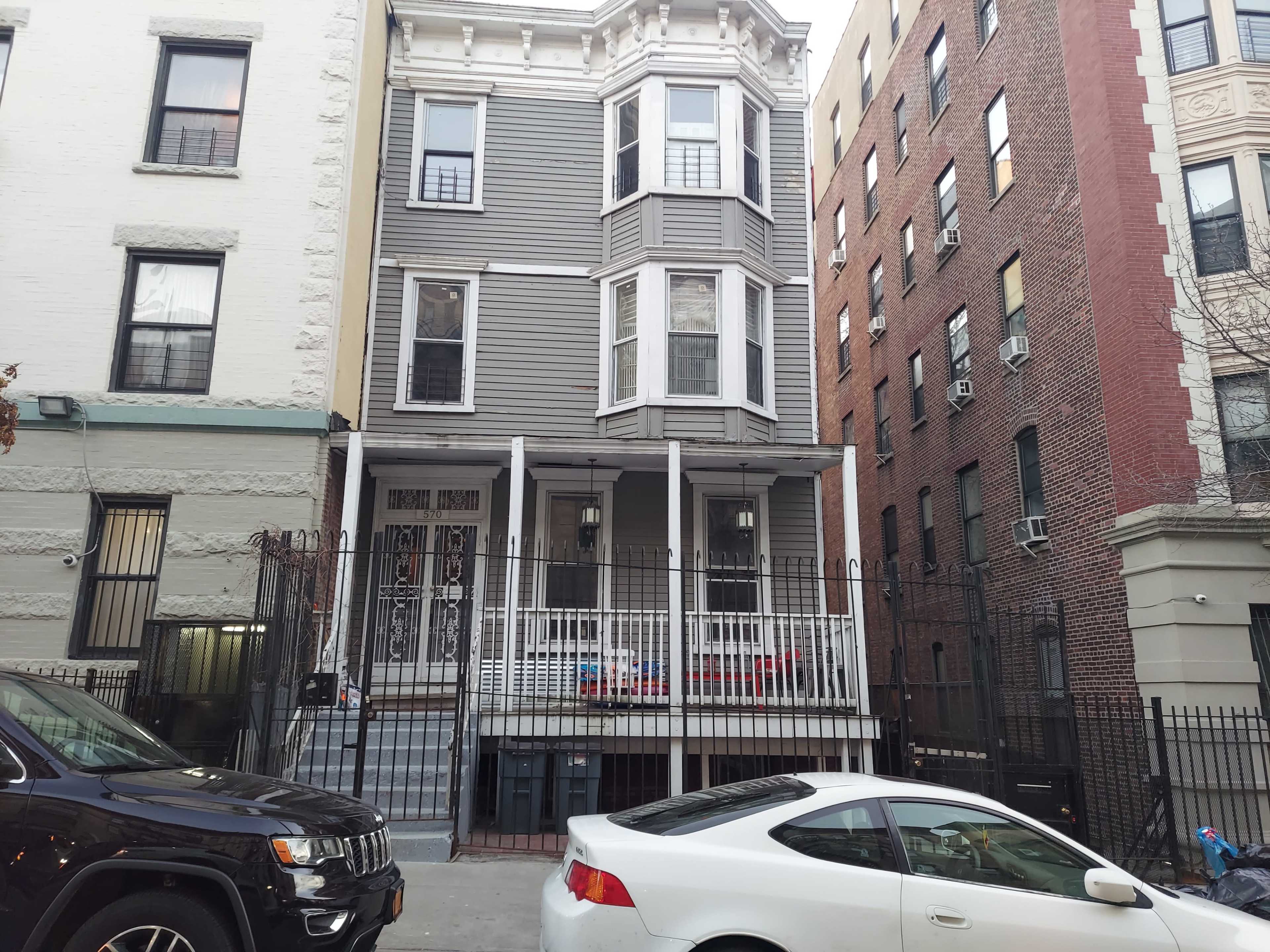 The image shows a three-story gray house with a front porch, flanked by two brick buildings on a city street.