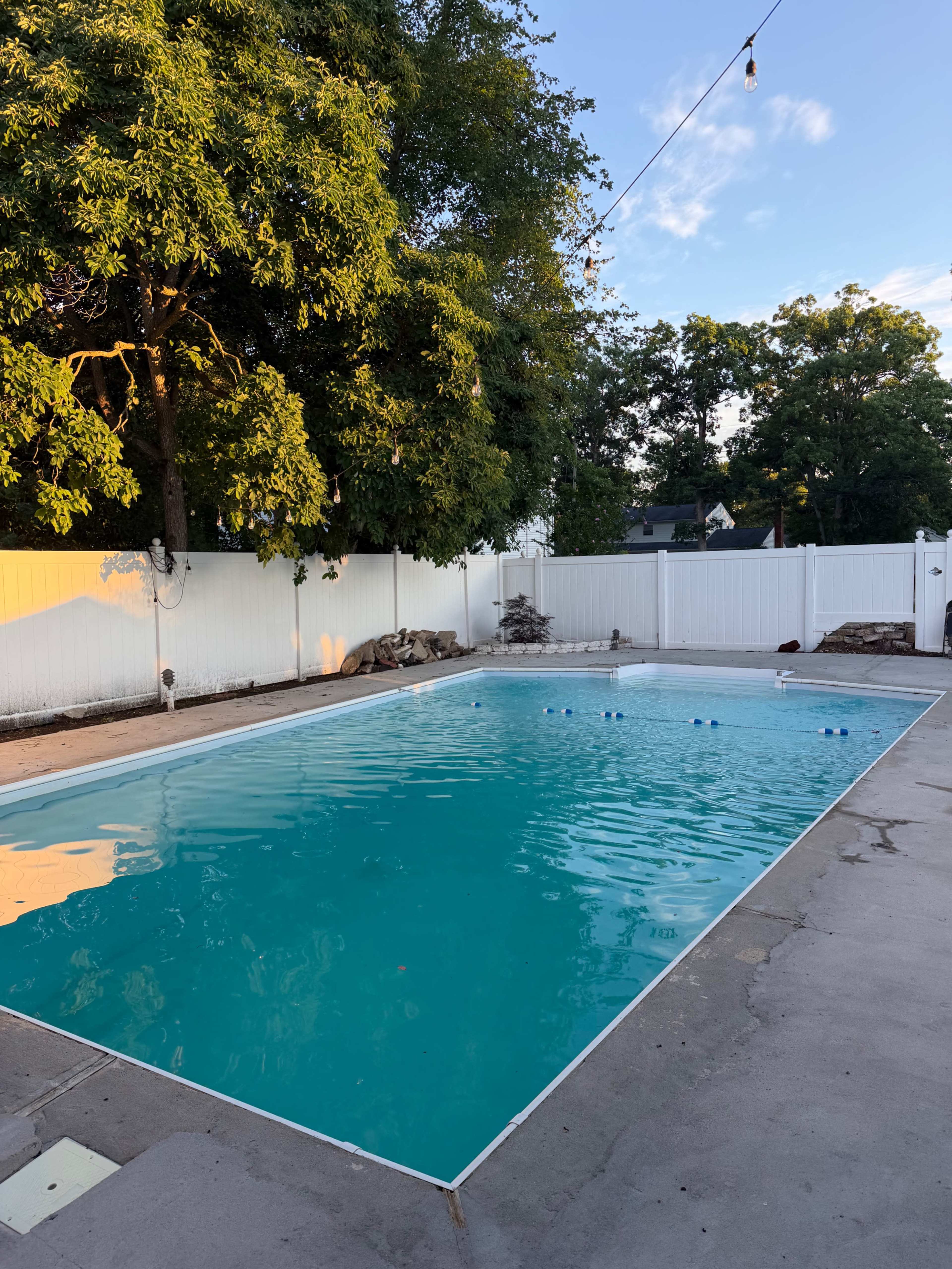 A clear swimming pool is surrounded by a white fence and trees, with decorative lights hanging above.