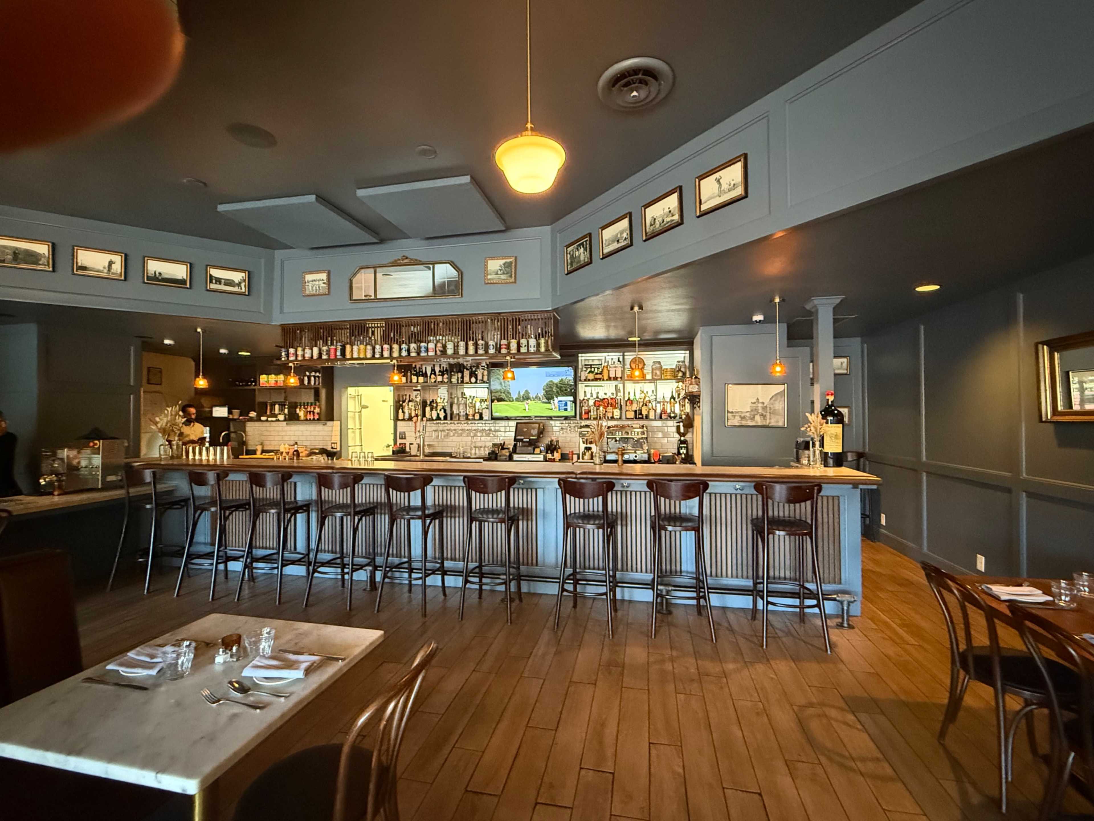 A well-lit bar area features a long counter with high stools, surrounded by shelves stocked with liquor and framed pictures on the walls.