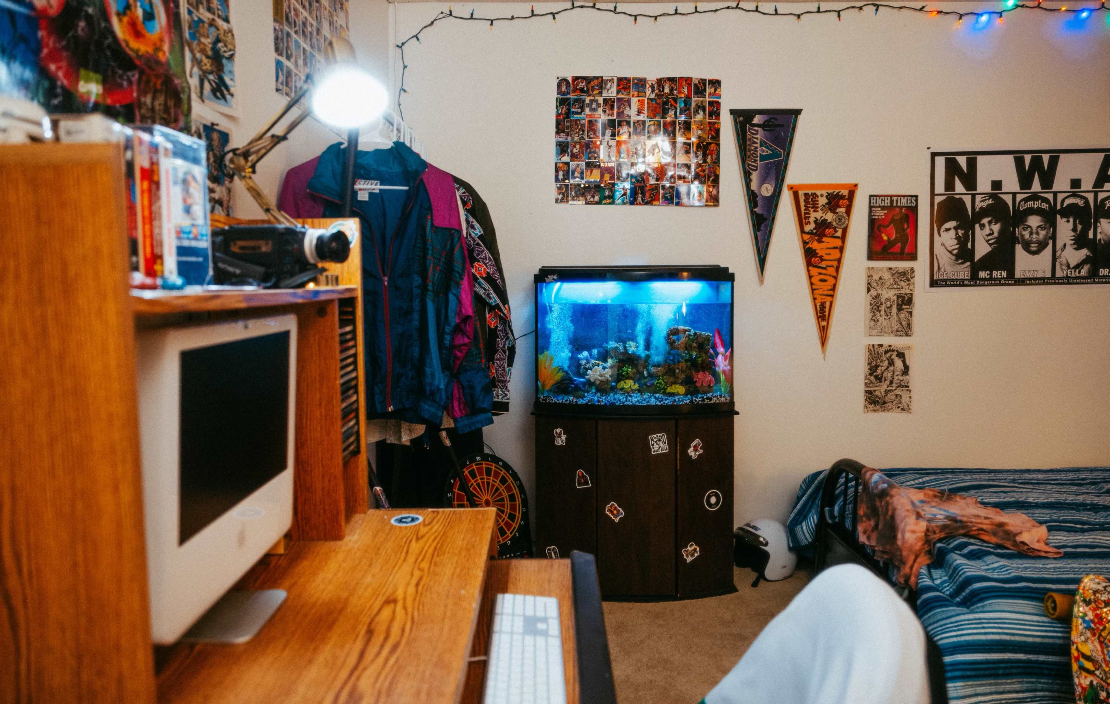 A colorful room featuring a computer desk, an aquarium, and a bed with a striped blanket, along with various posters and decorations on the walls.