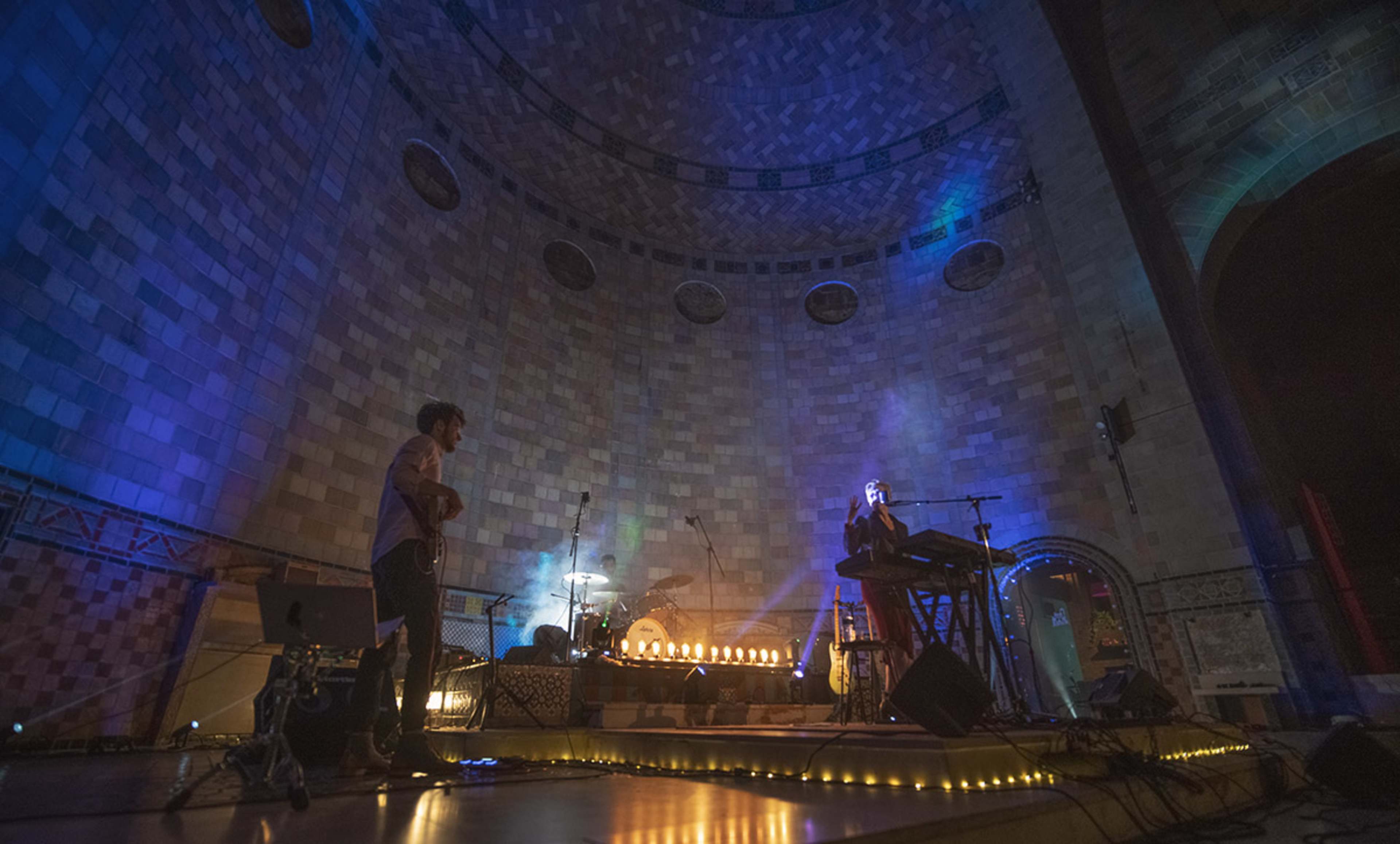 Two musicians perform on a stage with instruments and illuminated candles in a brick-walled venue.