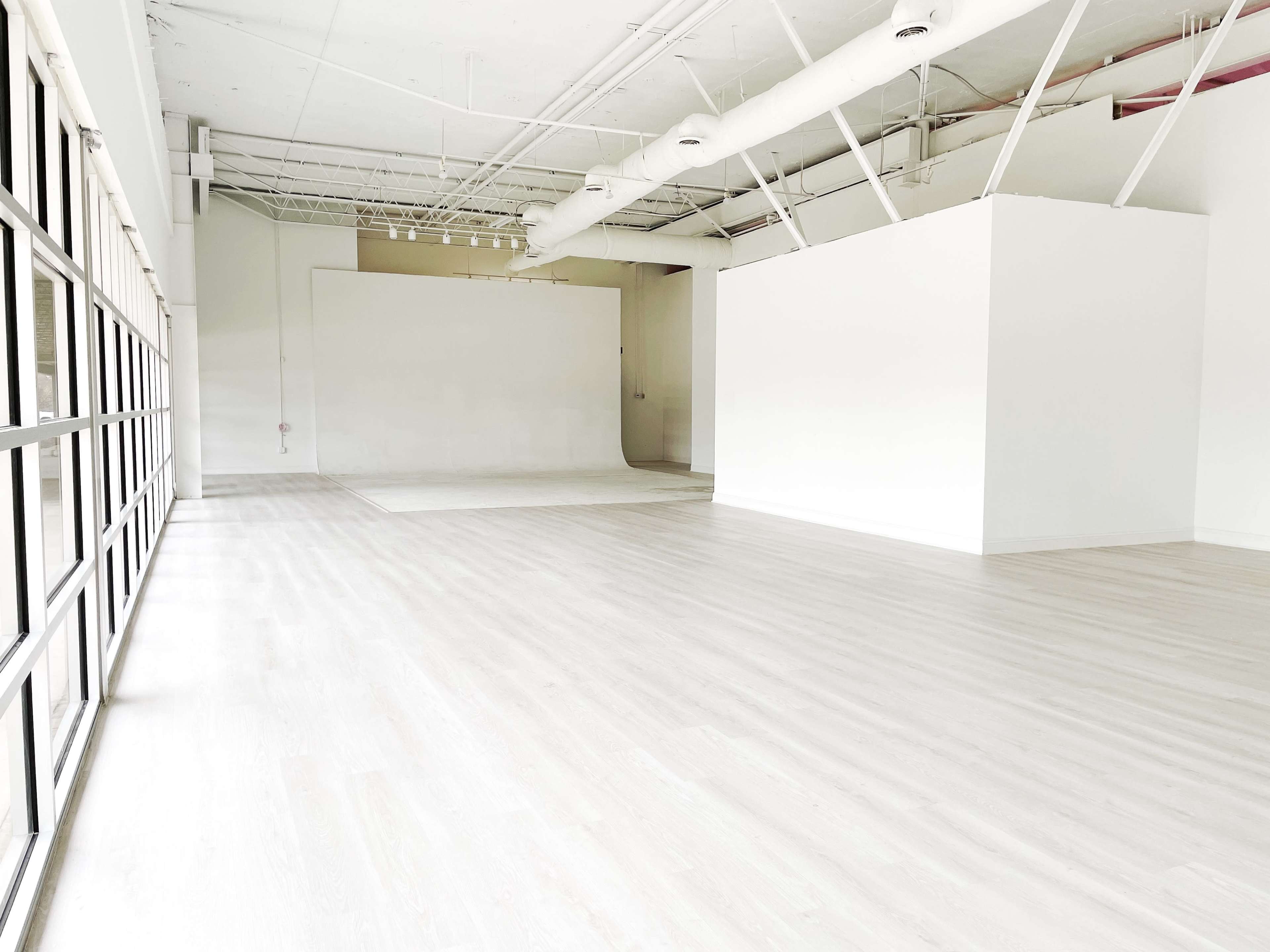 A spacious, empty room with light-colored flooring and white walls, featuring large windows along one side.