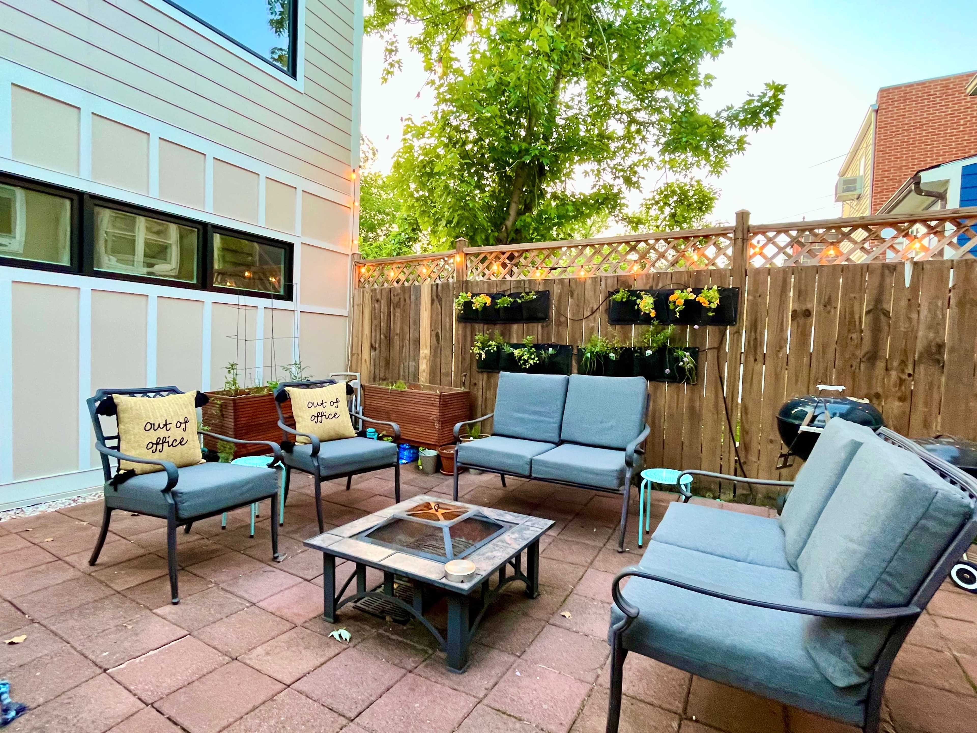 A patio area features a seating arrangement with gray chairs and a central fire pit, surrounded by a wooden fence with hanging plants.