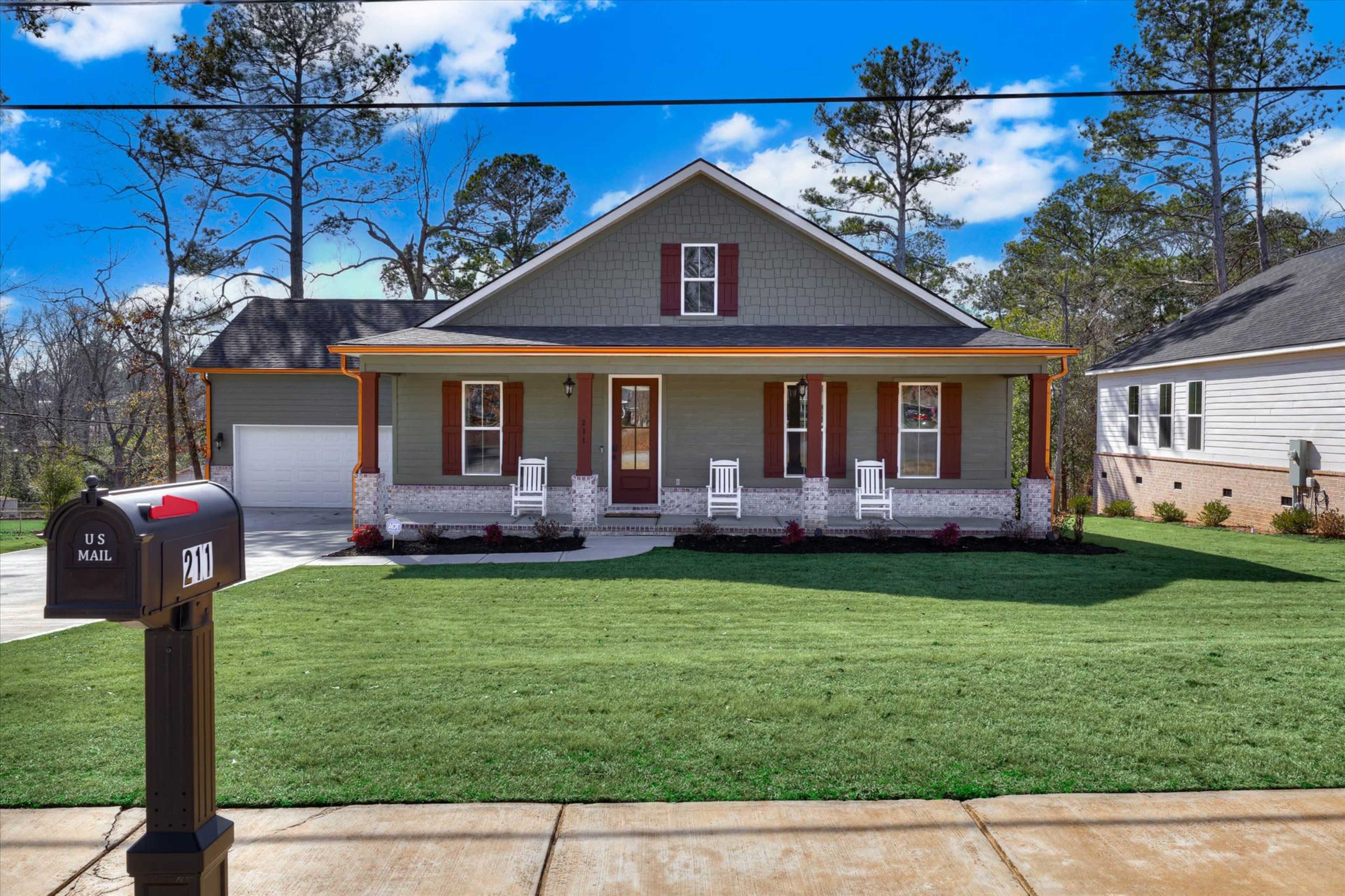 A single-story house with a gray exterior, red shutters, and a front porch featuring white rocking chairs is set in a landscaped yard with freshly mowed grass.
