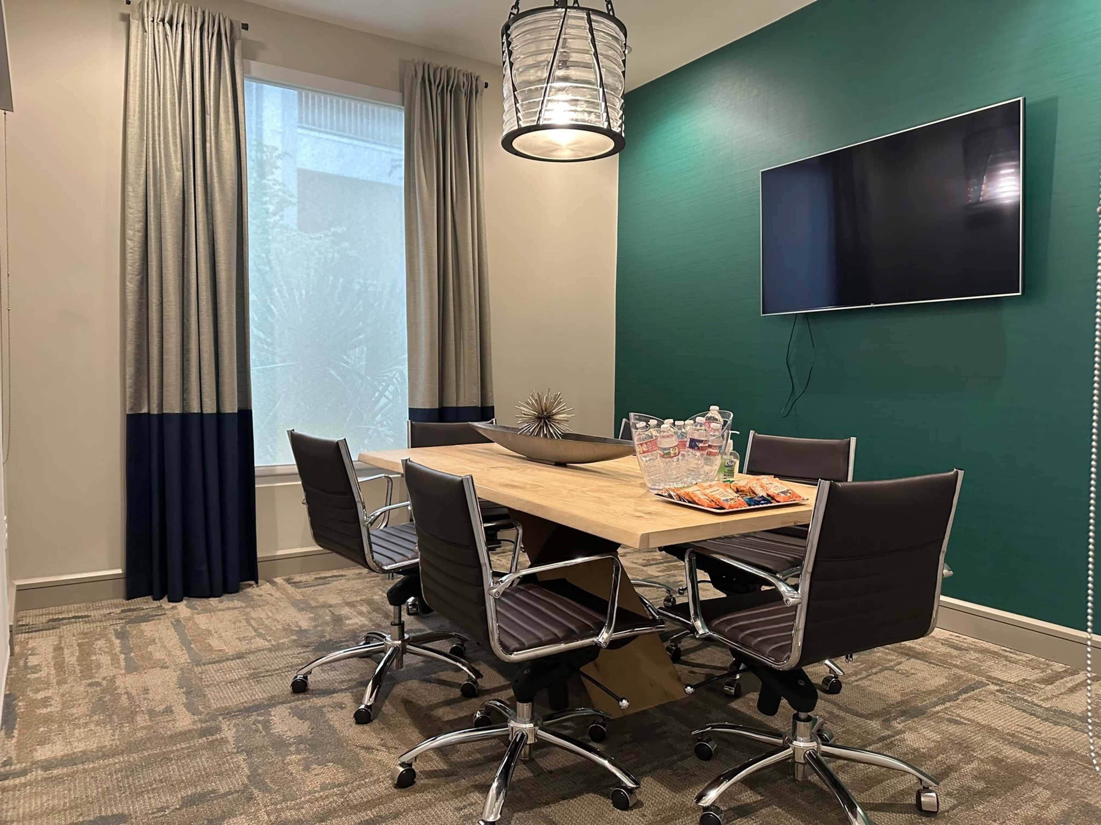 A conference room features a wooden table surrounded by six chairs, a television mounted on the wall, and curtains framing a window.