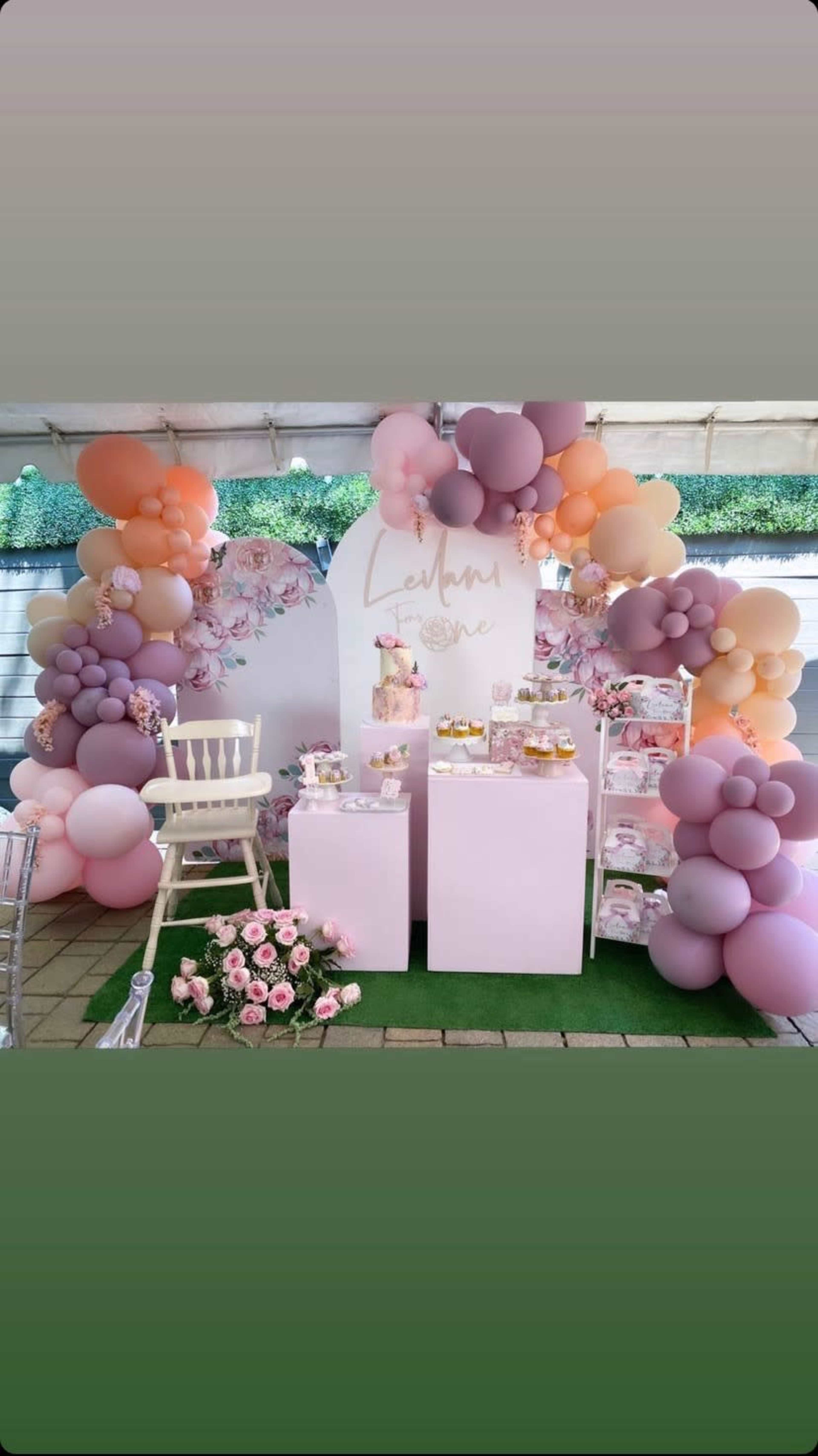 A decorated event space featuring a backdrop with balloons and floral arrangements, alongside dessert tables with a cake and treats.