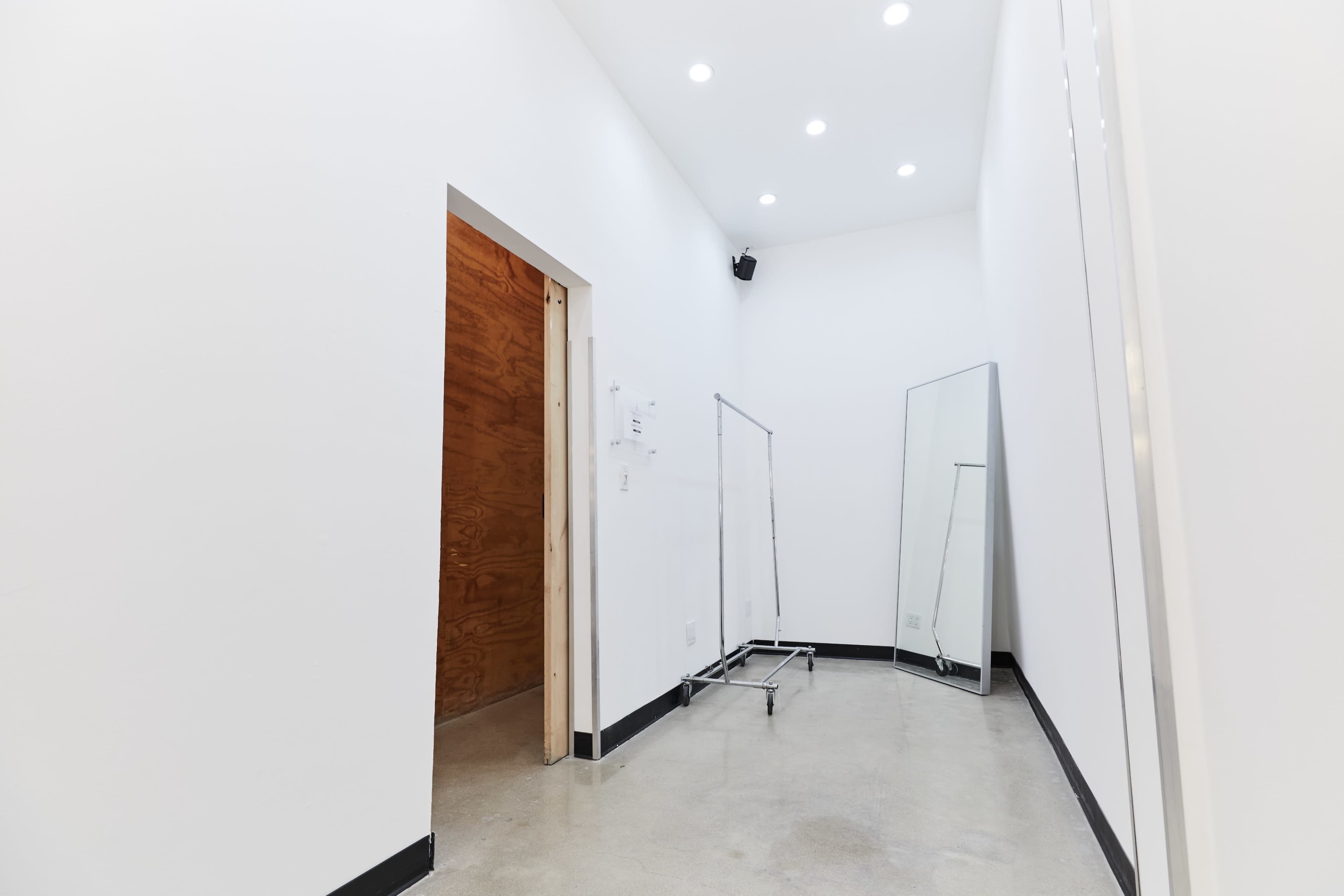 The image shows a small, empty room with white walls, a wooden door on one side, a clothing rack, and a full-length mirror.