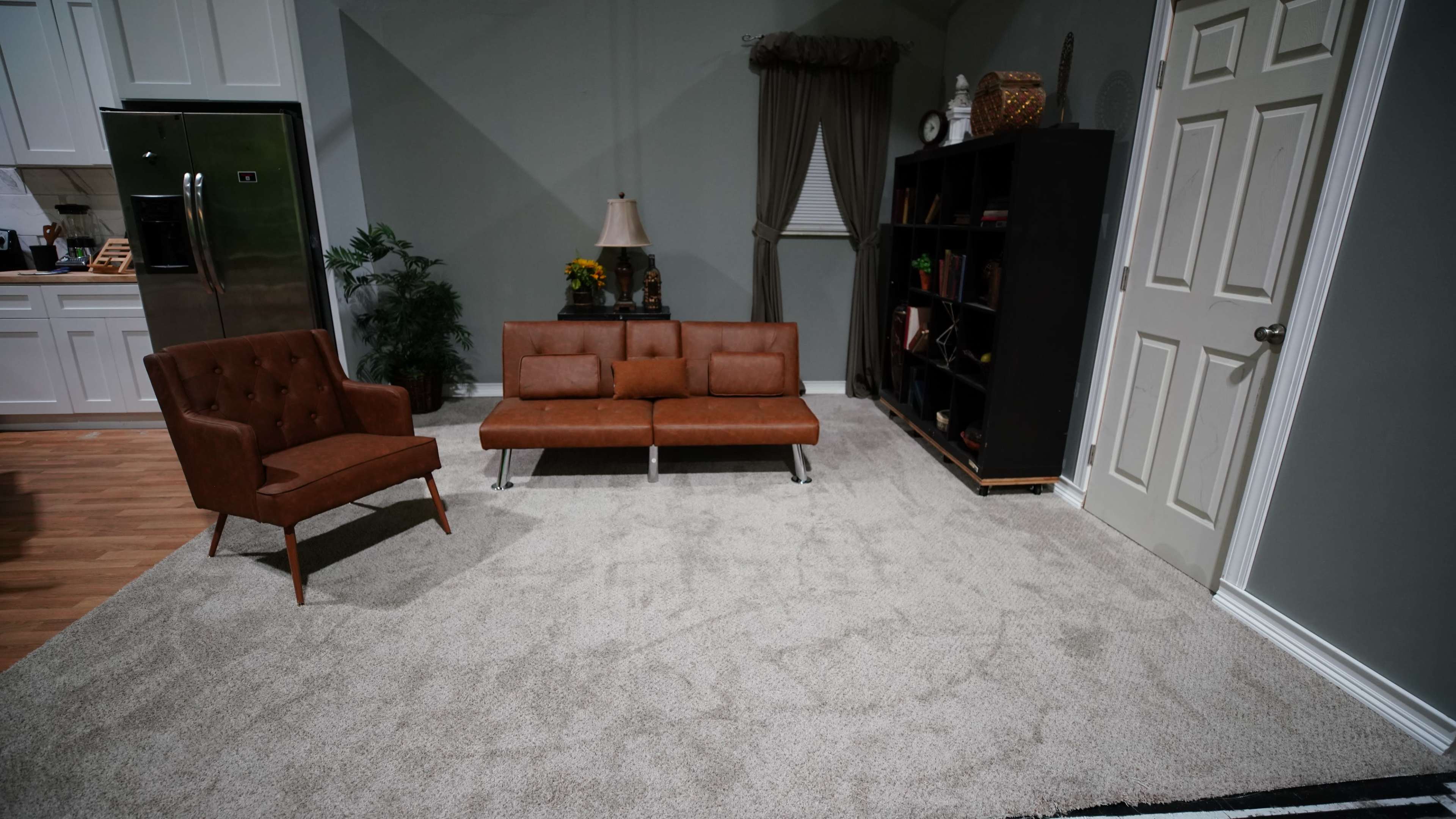The scene shows a living room with a brown leather couch, an orange armchair, a bookshelf, and a light-colored carpet.