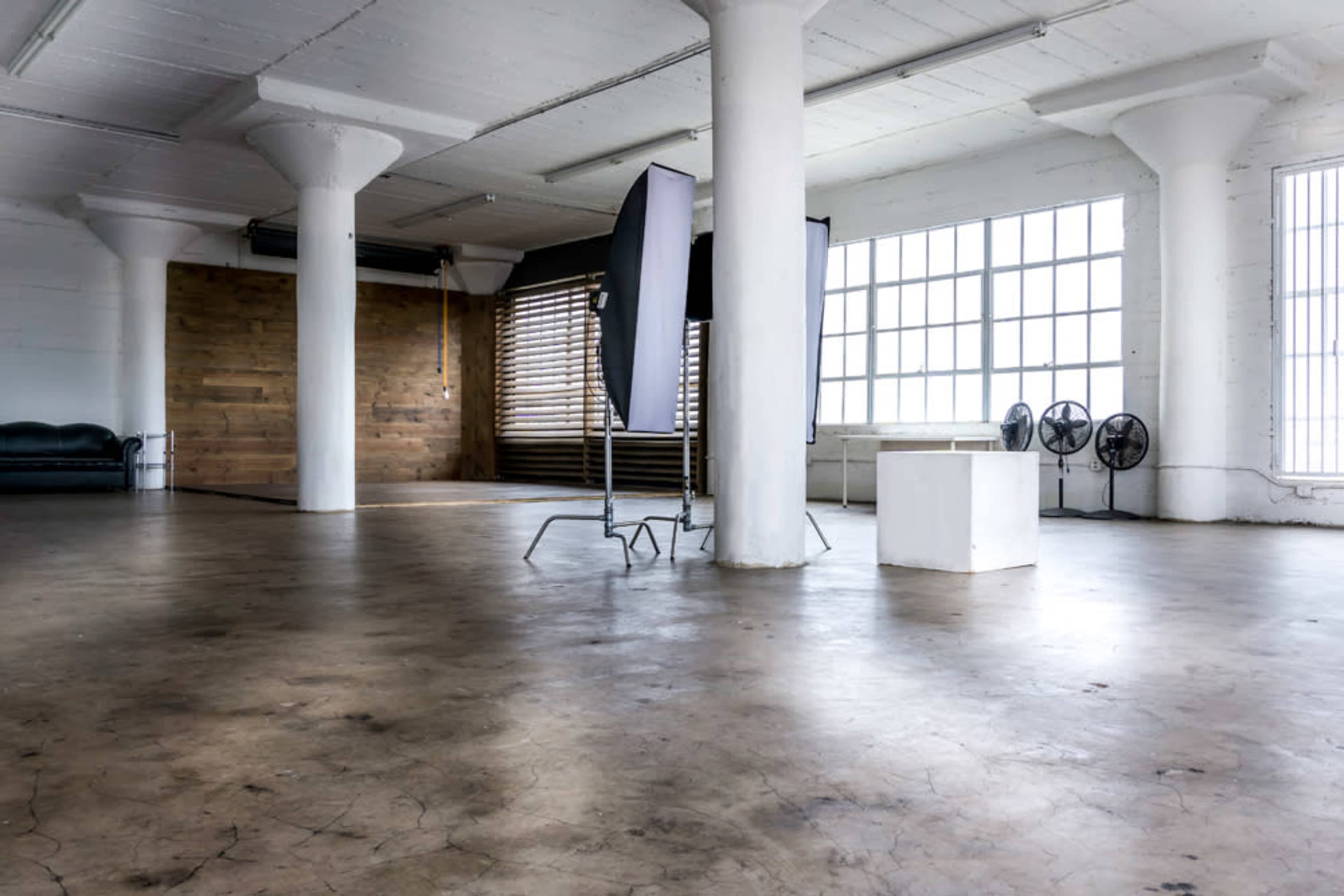The image shows a spacious, well-lit studio with concrete floors, large windows, and photo equipment set up in the center.