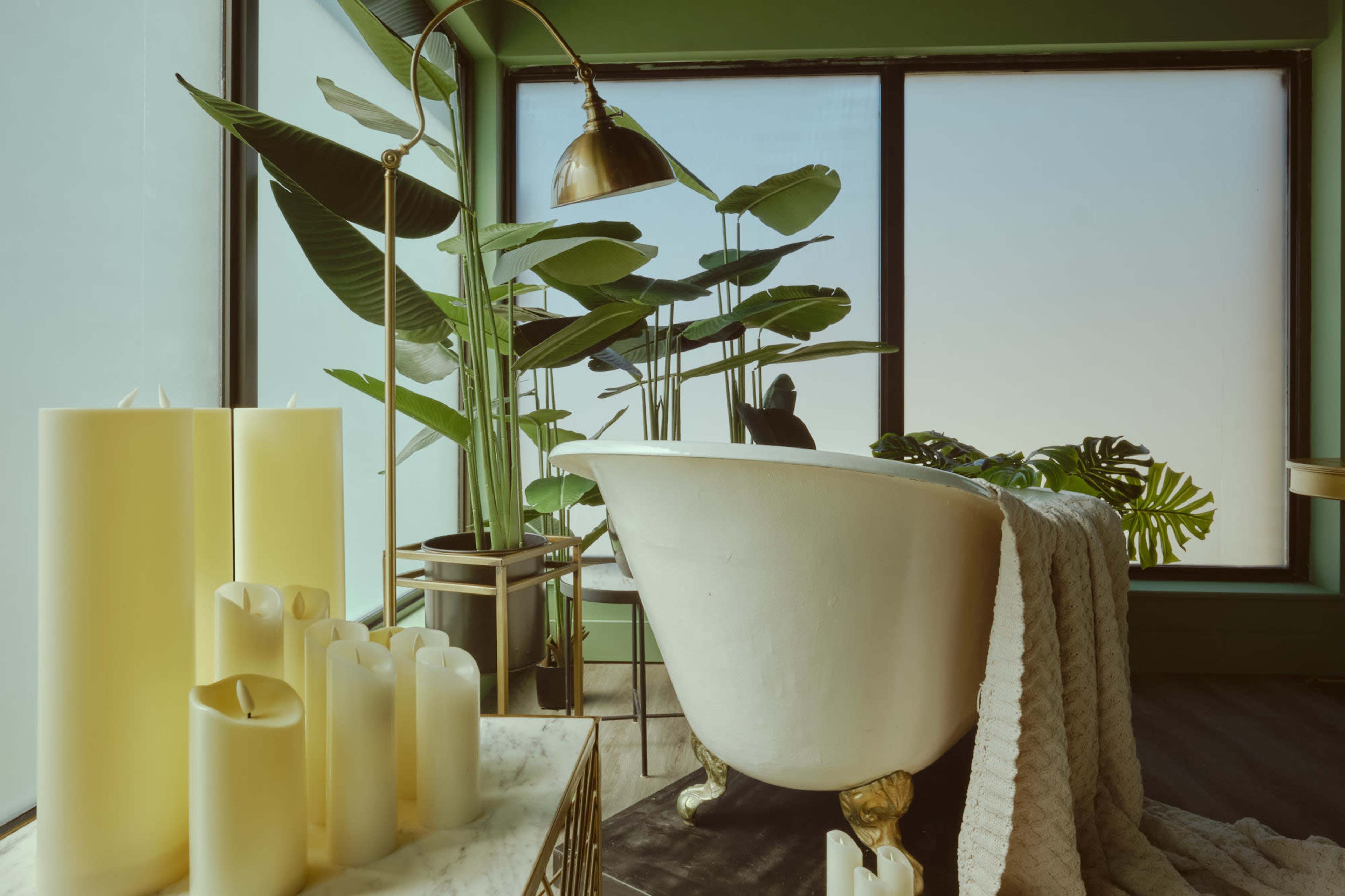A stylish bathroom features a freestanding bathtub beside large windows and greenery, with decorative candles on a marble surface.