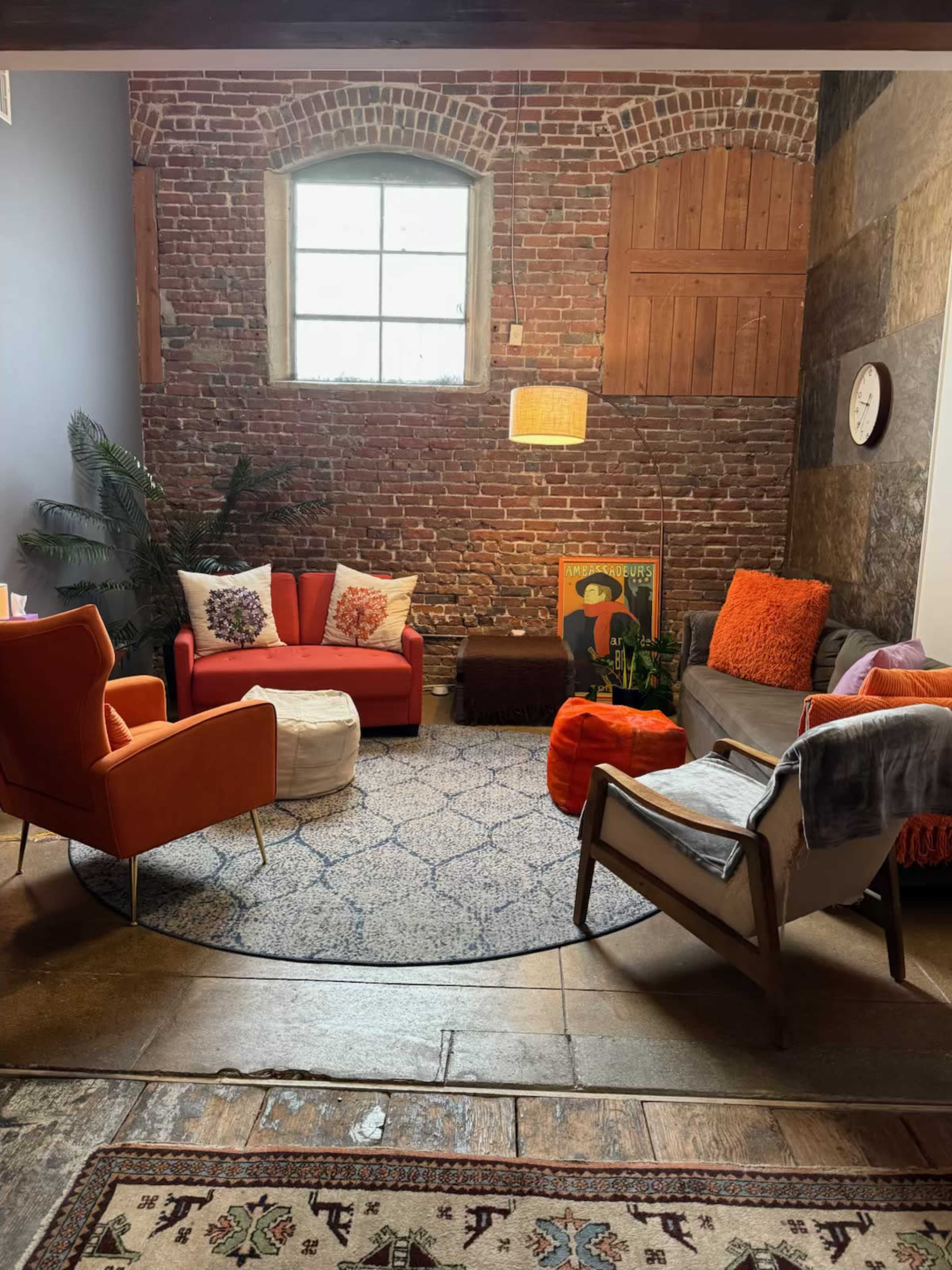 The image shows a cozy living space featuring a mix of colorful chairs, a round rug, a coffee table, and a large brick wall with a window.