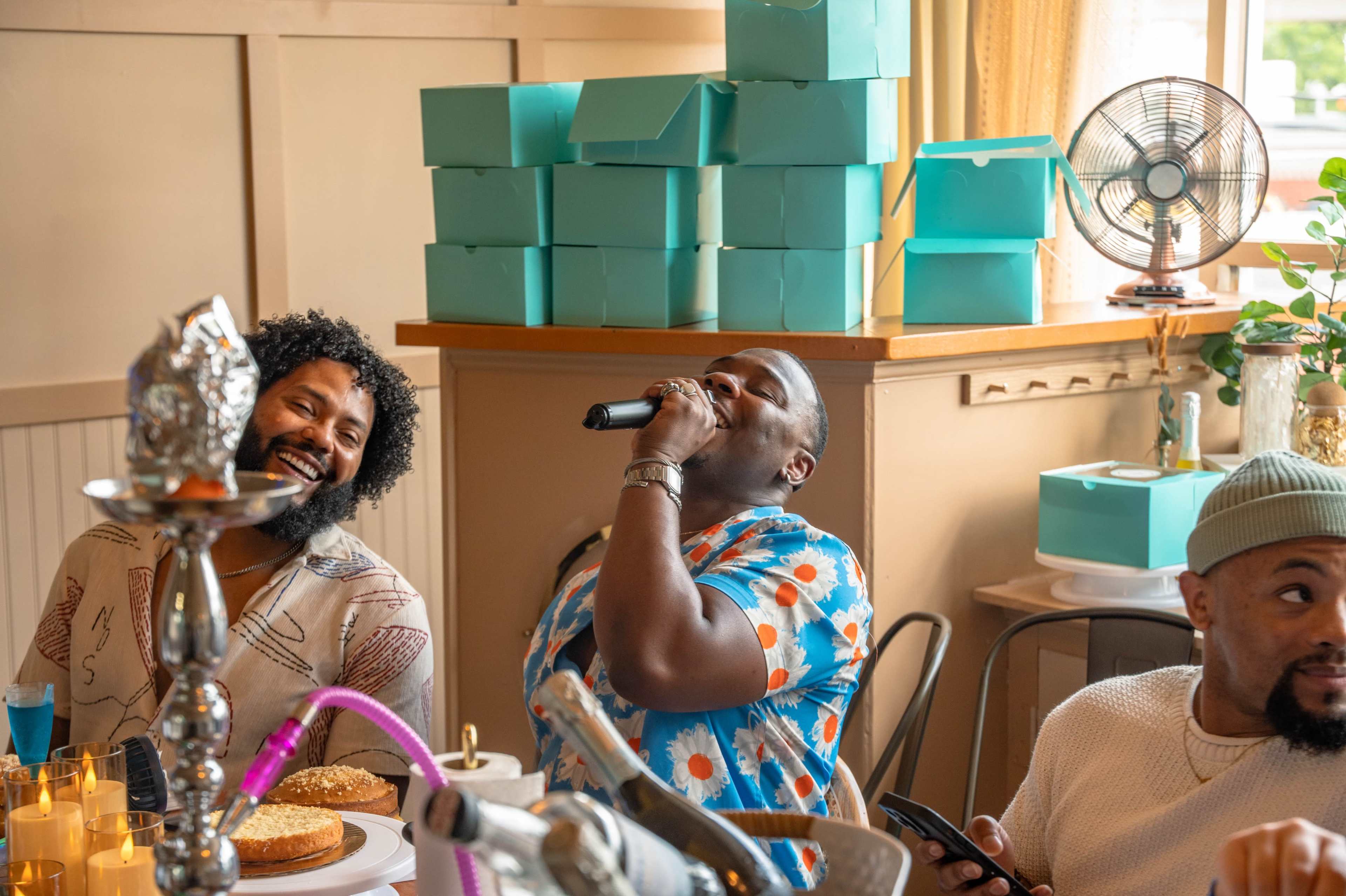 A group of friends enjoys a gathering, with one person singing into a microphone while others laugh and interact in a bright room filled with teal boxes.