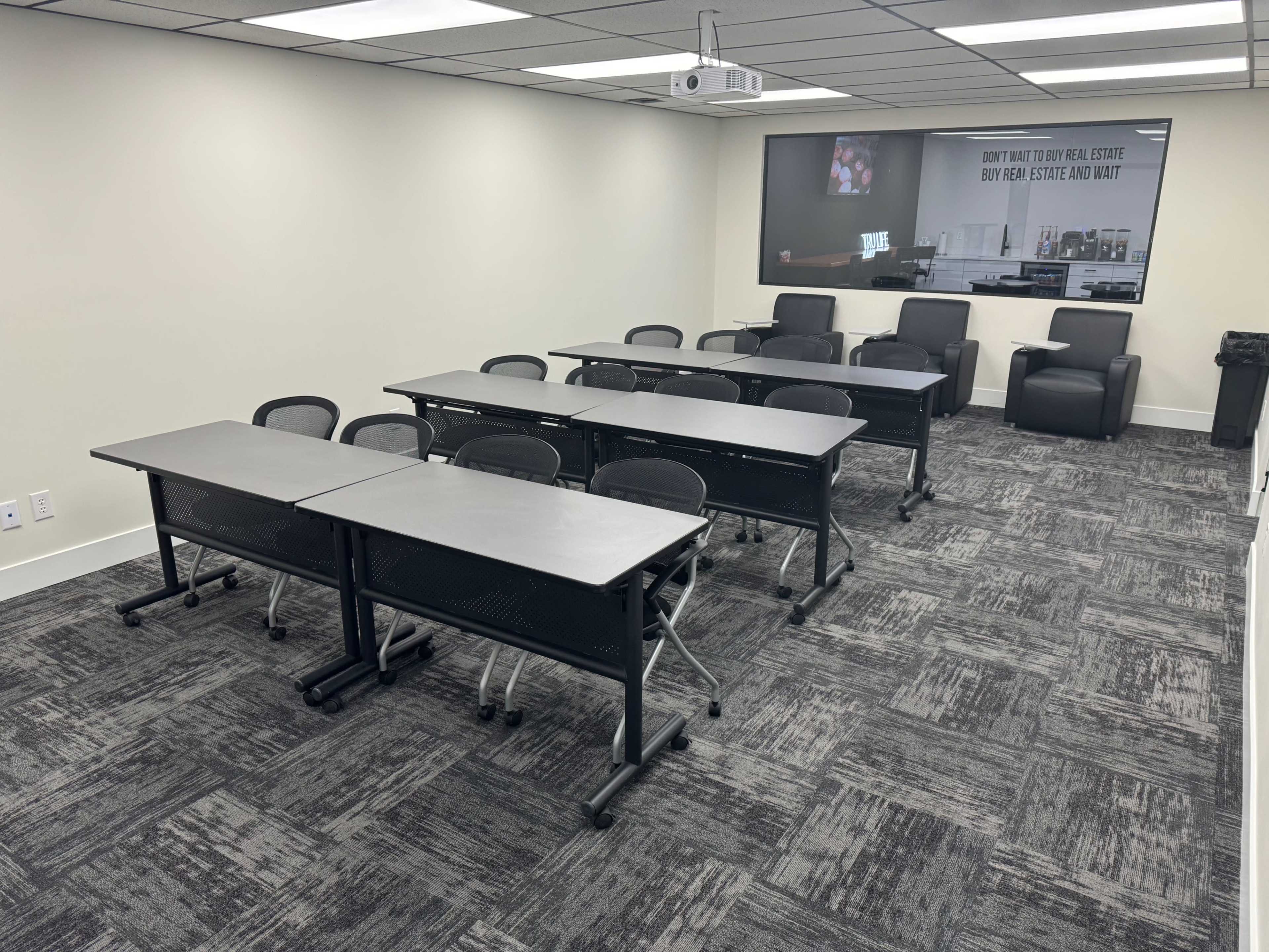 A modern classroom or meeting room set up with multiple tables and chairs arranged in rows, a projection screen on the wall, and a couple of armchairs in the corner.