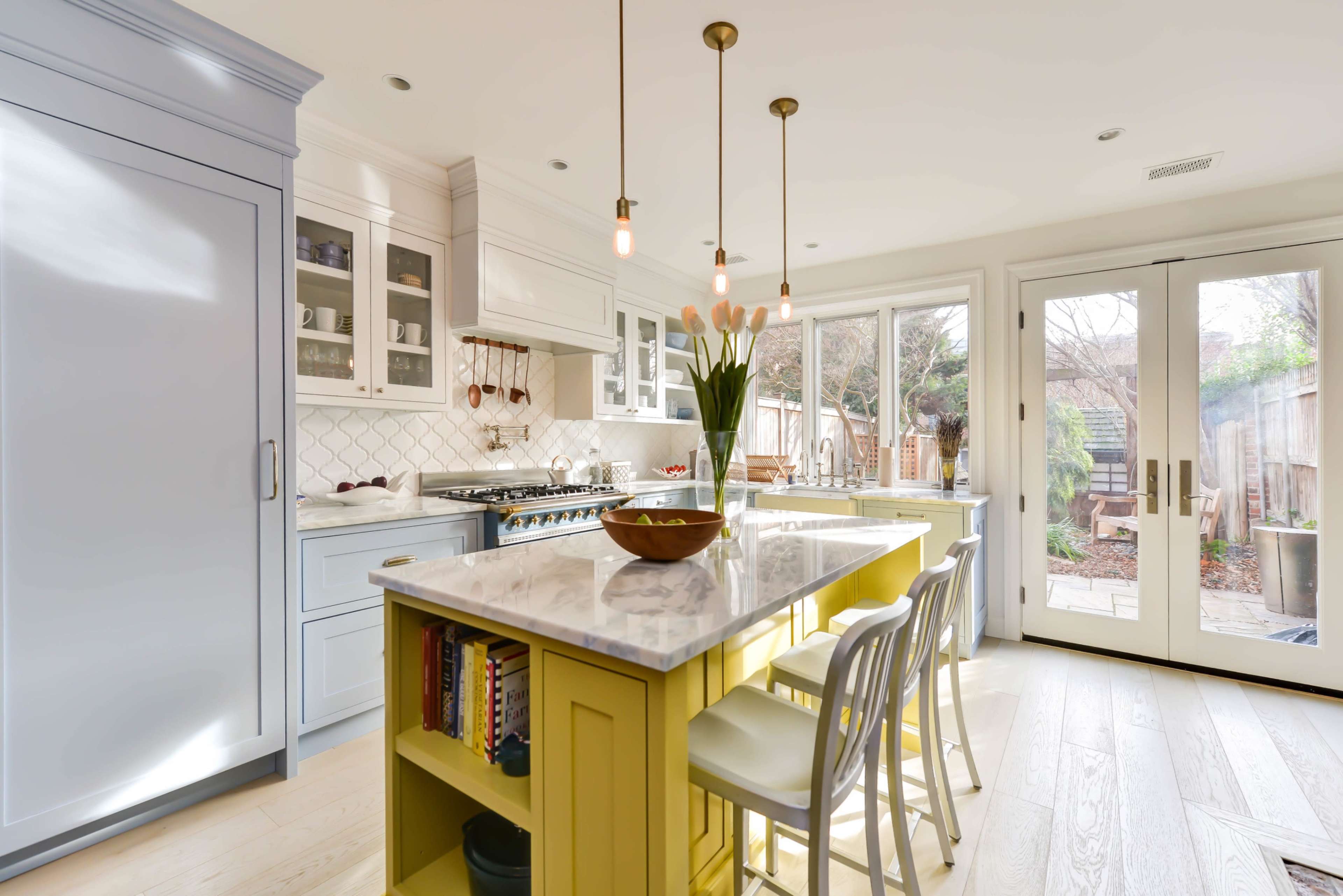 A modern kitchen with light-colored cabinetry, a large island featuring a yellow base, and natural light streaming in through windows and doors.