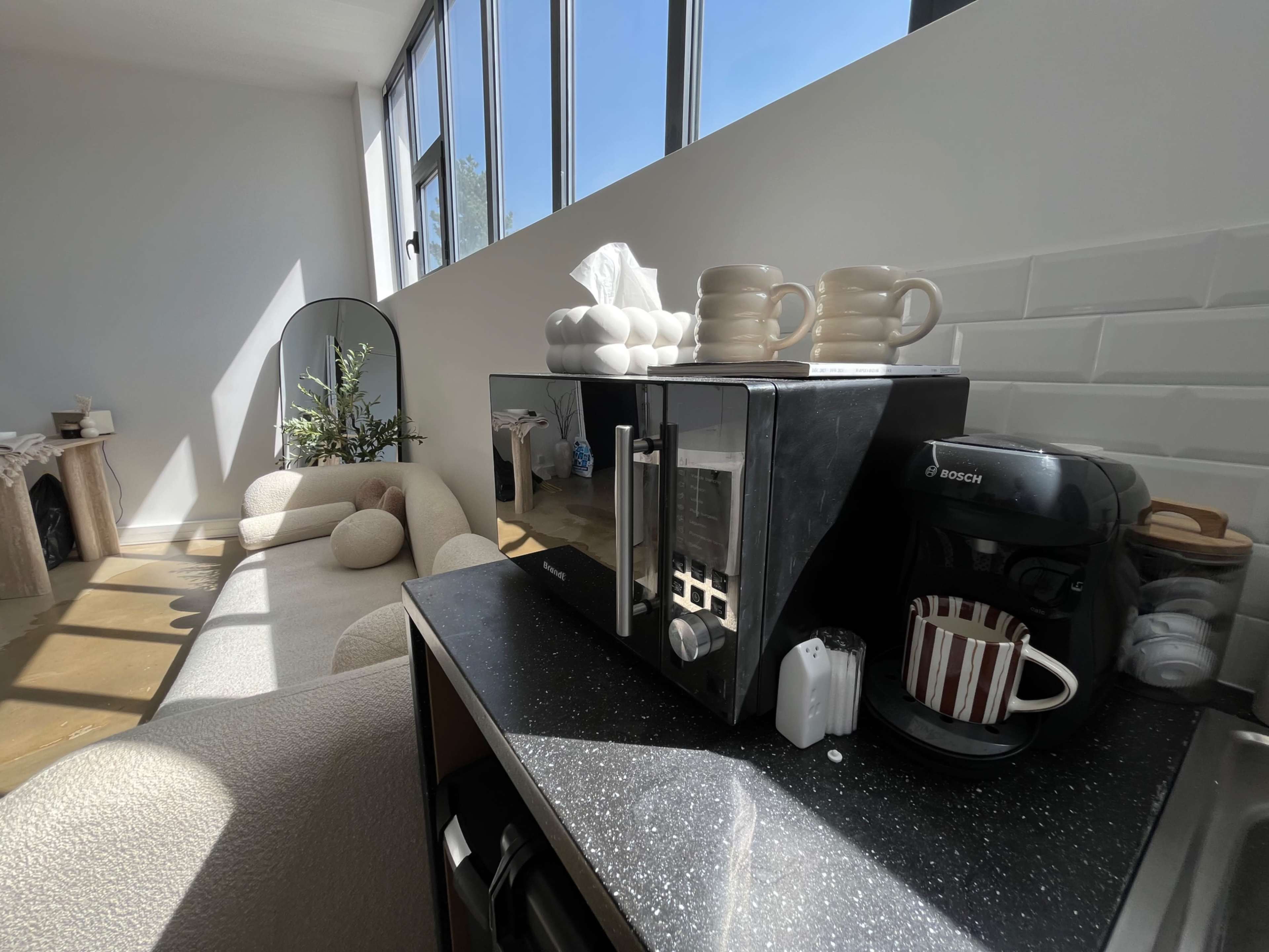 The image shows a modern kitchen space featuring a microwave, a coffee maker, and a cozy sofa, with ample natural light streaming through large windows.