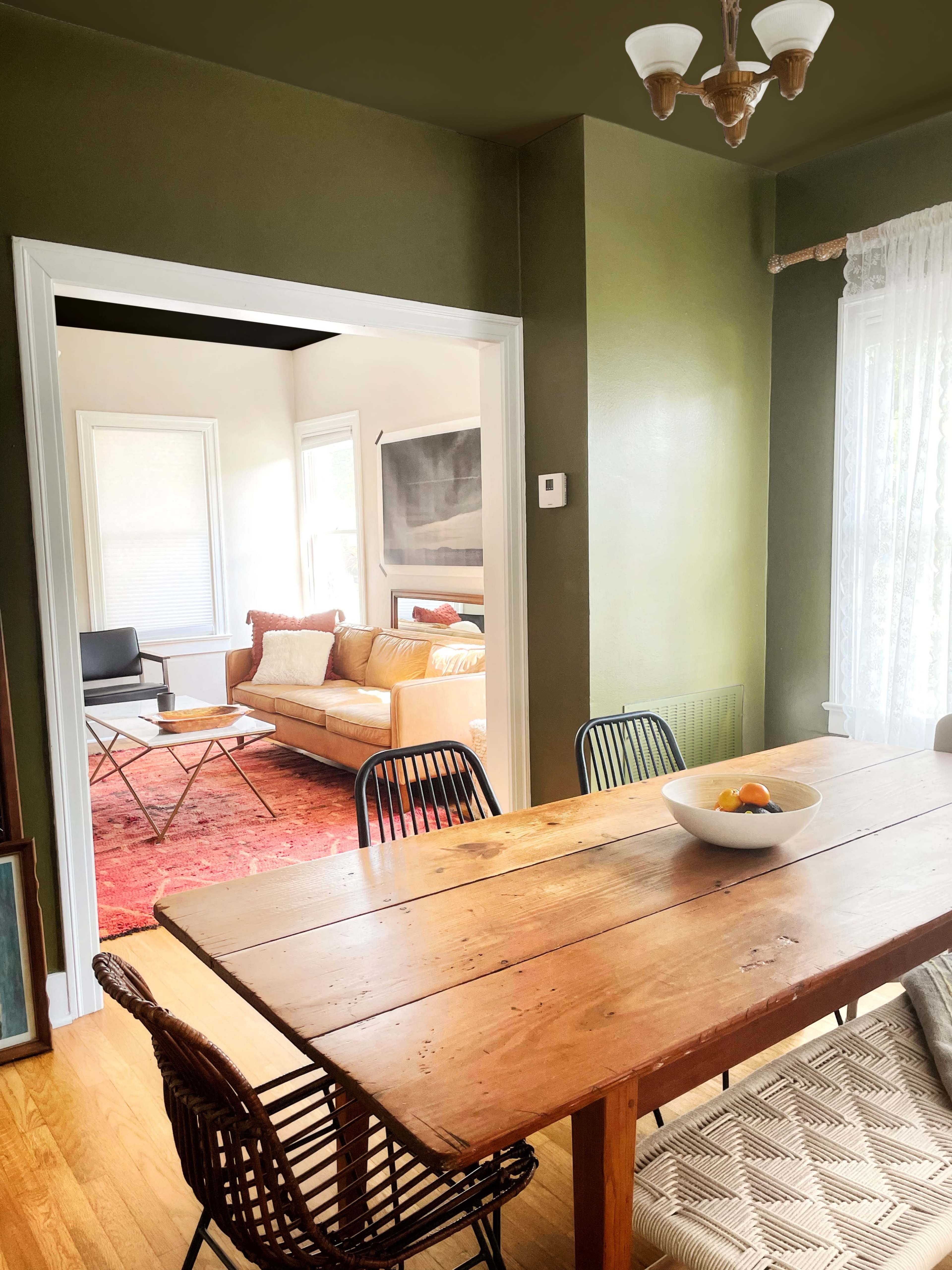 A wooden dining table with black chairs is set in a green-walled room, which opens to a bright living area featuring a couch and a television.