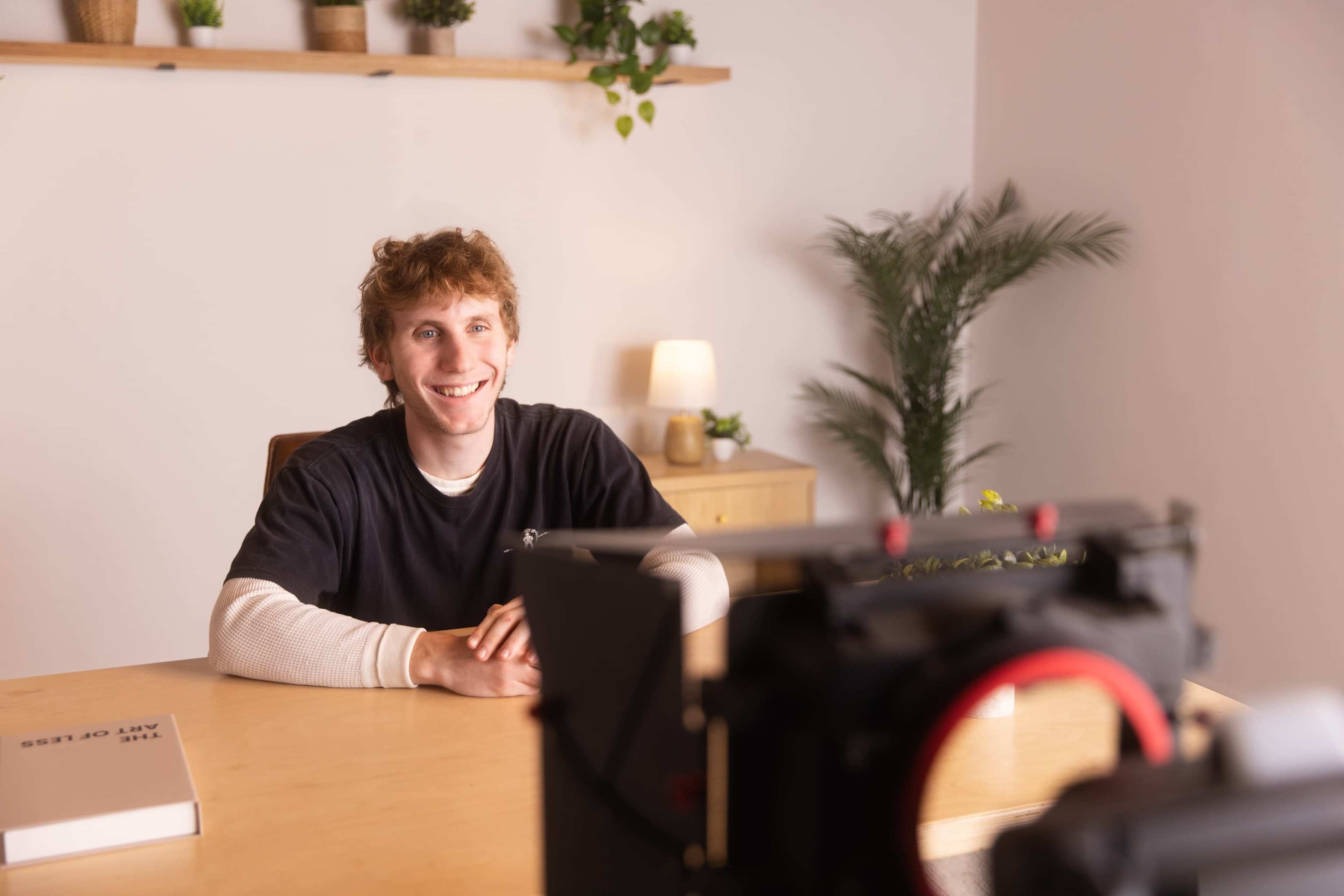 A person sits at a wooden table in a well-lit room, facing a camera while smiling.