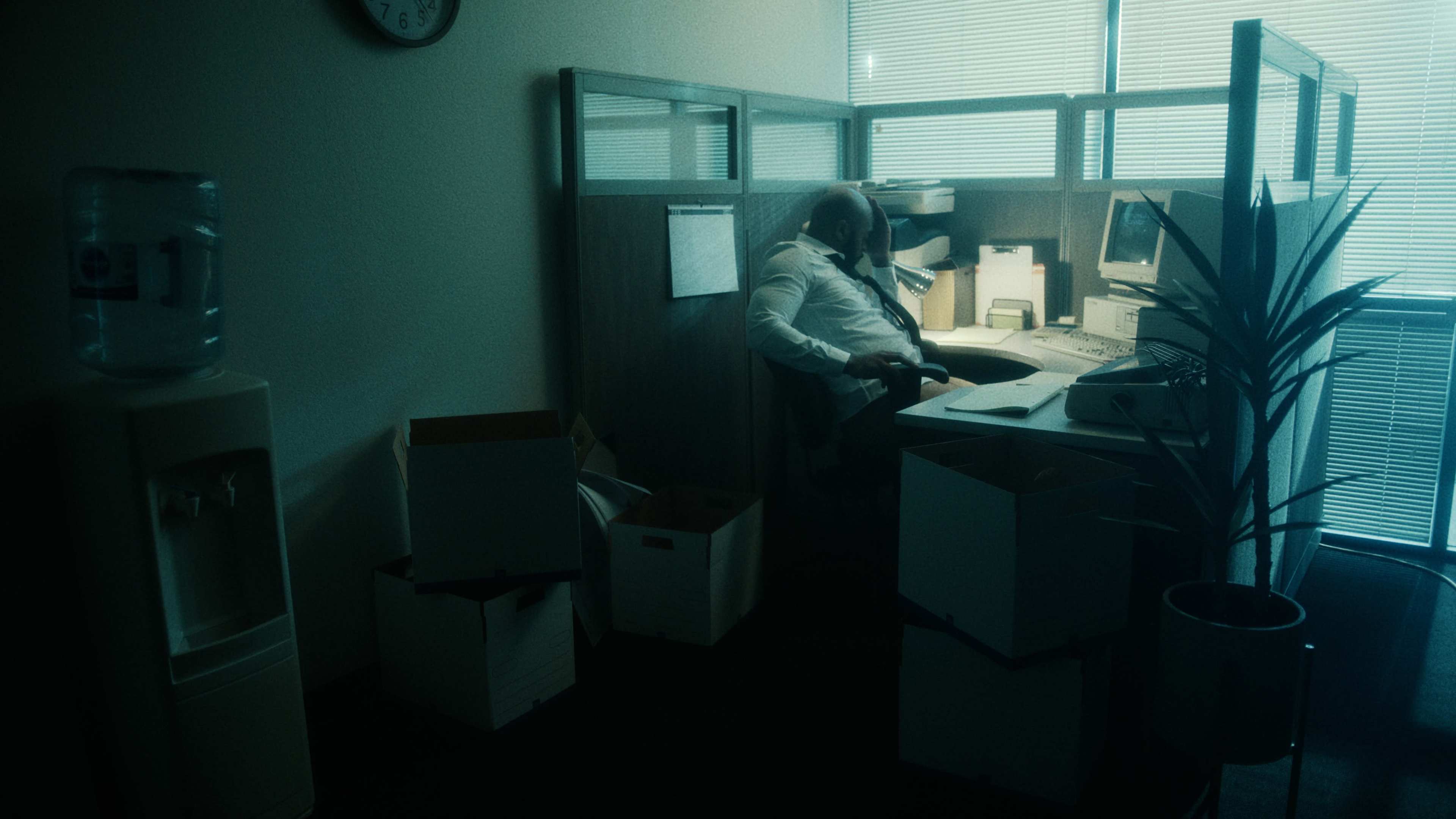 An office cubicle is dimly lit, featuring a seated man at a cluttered desk surrounded by cardboard boxes.
