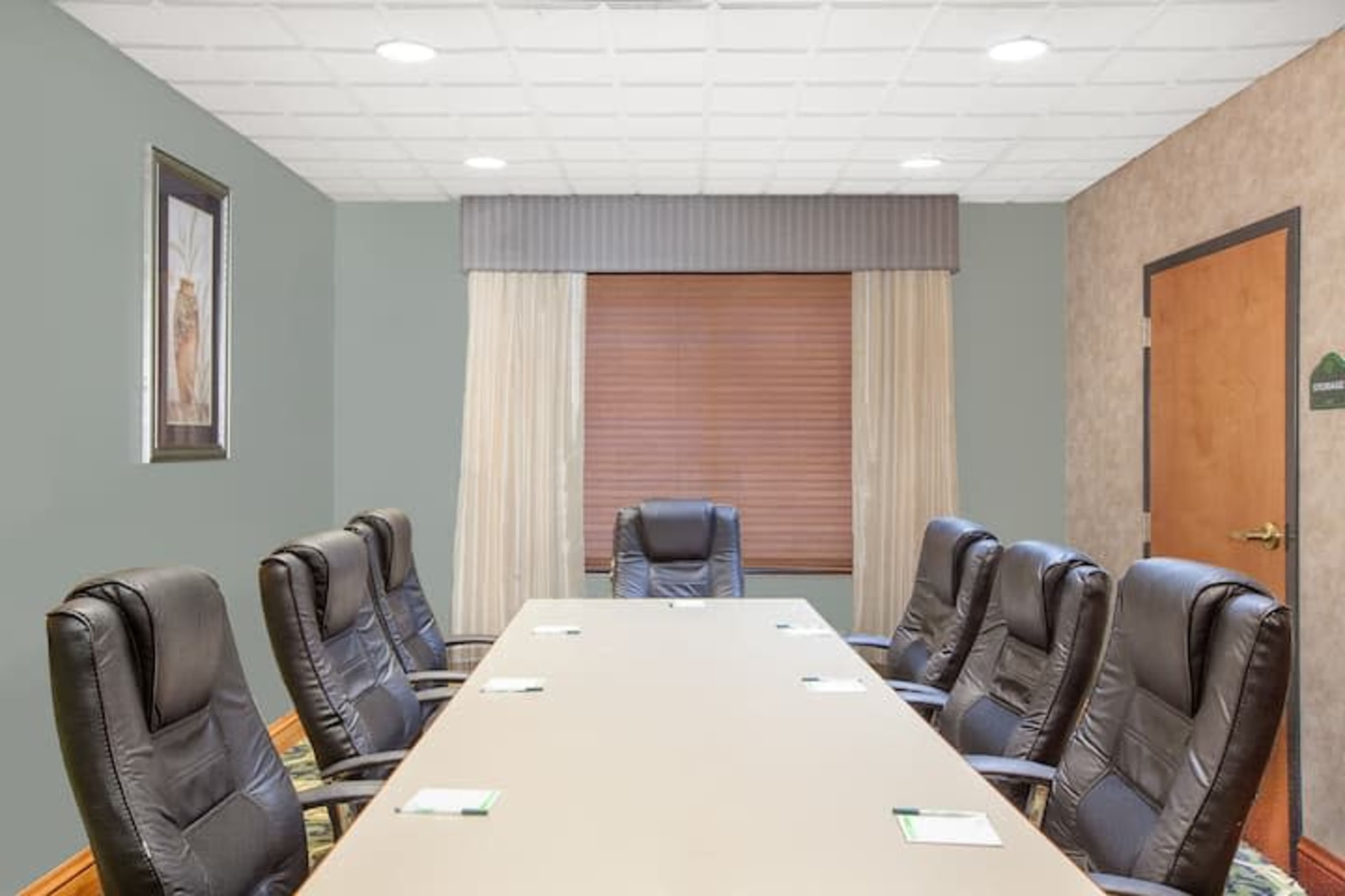 A conference room features a long table surrounded by black swivel chairs under overhead lights, with a window covered by blinds and curtains.