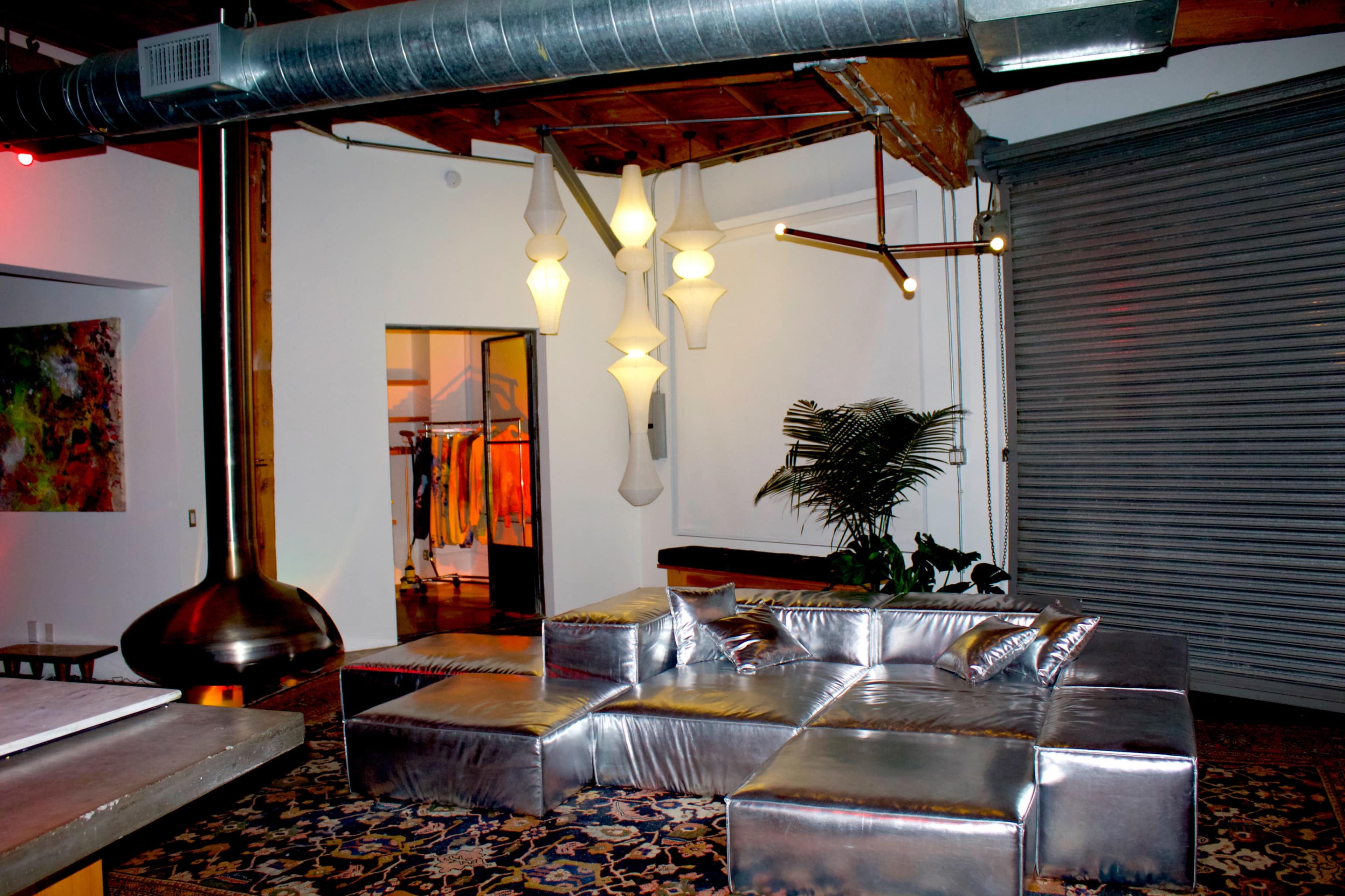 The image shows a modern living space featuring a shiny silver sectional sofa, unique lighting fixtures hanging from the ceiling, and a large plant beside a closed rolling garage door.