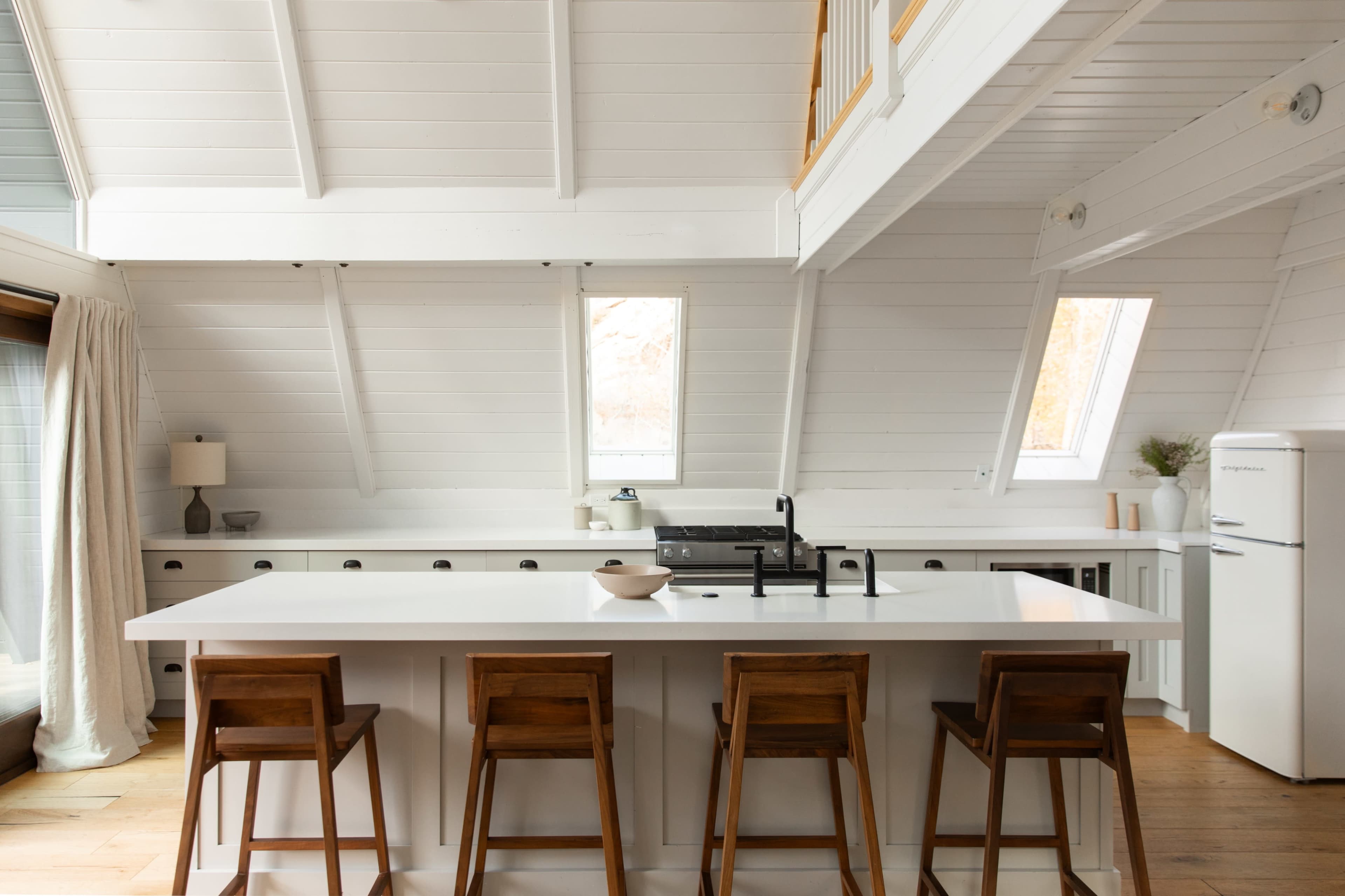 A modern kitchen with a white color scheme, featuring an island with four wooden bar stools and large windows allowing natural light.