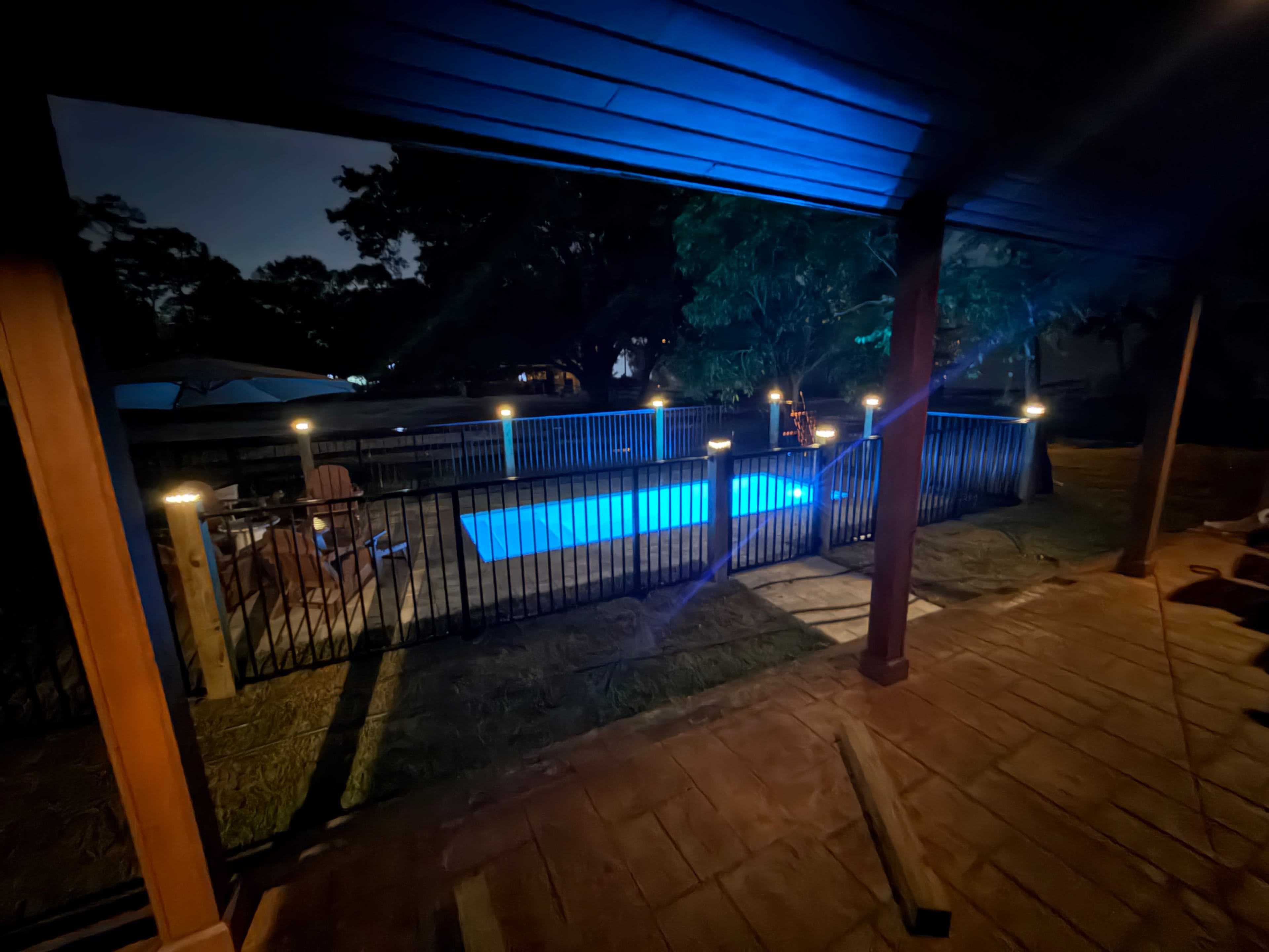 The image shows a well-lit swimming pool surrounded by a fence, viewed from a covered patio at night.
