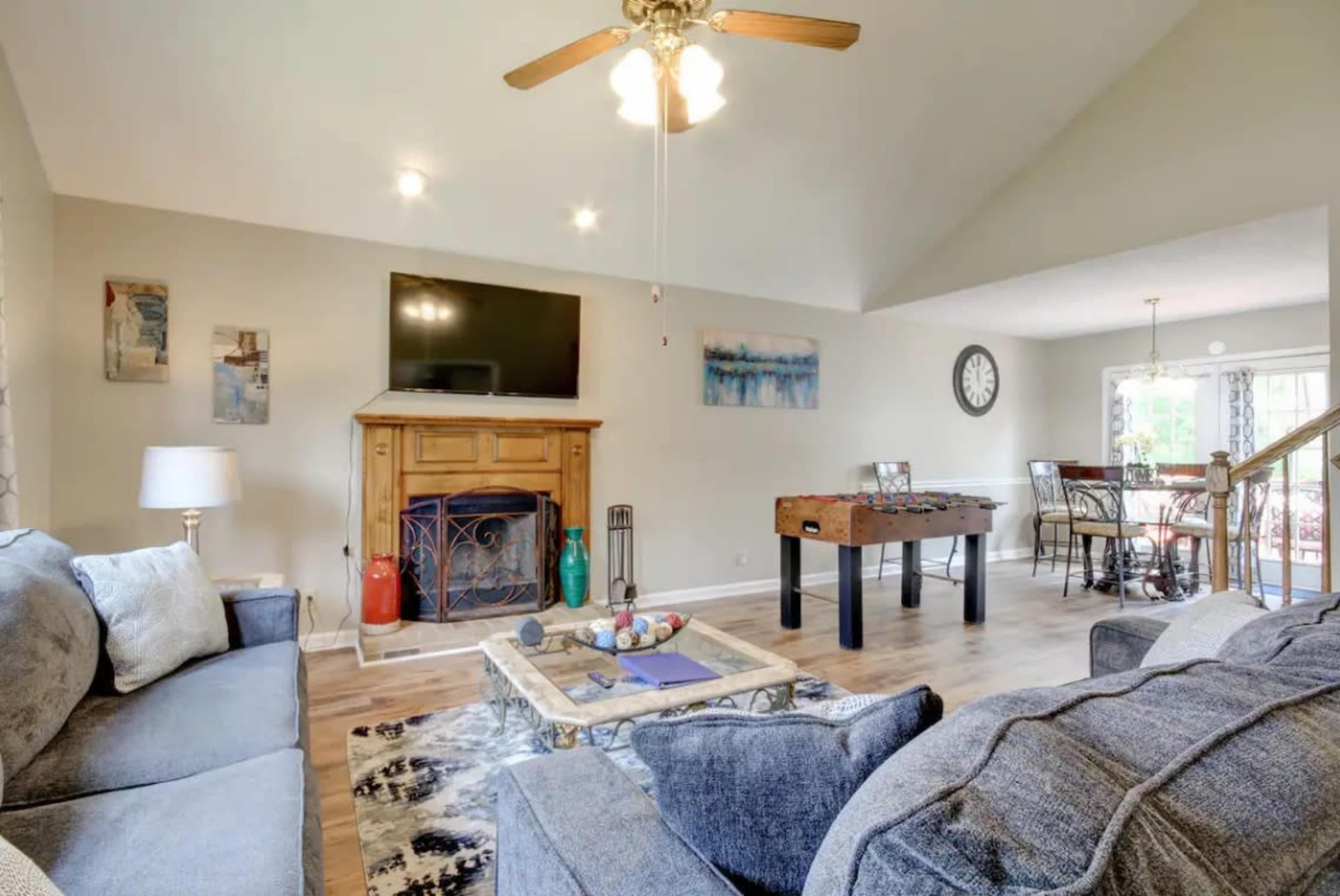 The living room features a ceiling fan, a wooden fireplace with a television mounted above it, and a foosball table, with seating arrangements and decorative elements throughout.