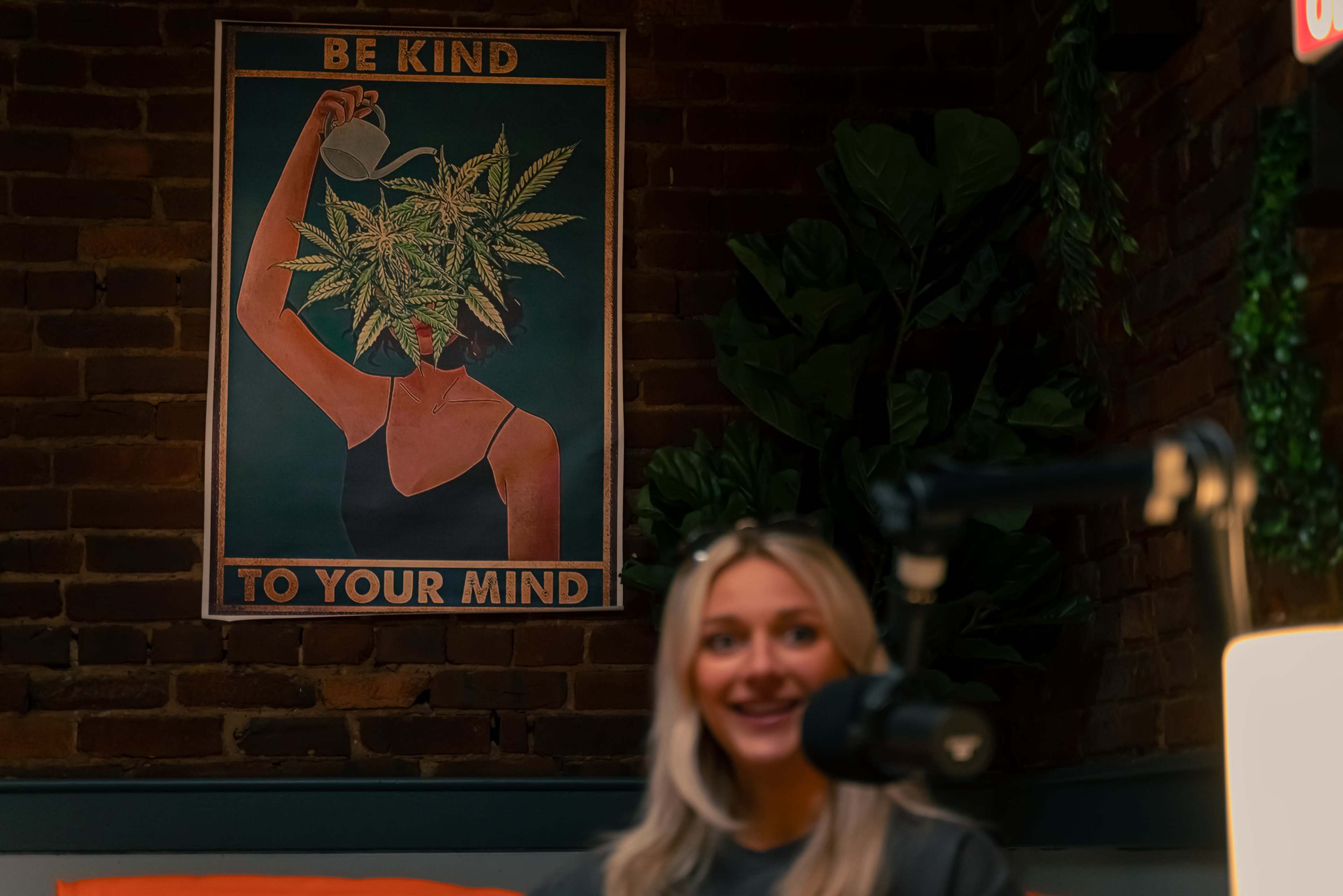 A woman with blonde hair sitting in front of a colorful poster that promotes kindness and mental well-being, illustrated with a person watering a cannabis plant.