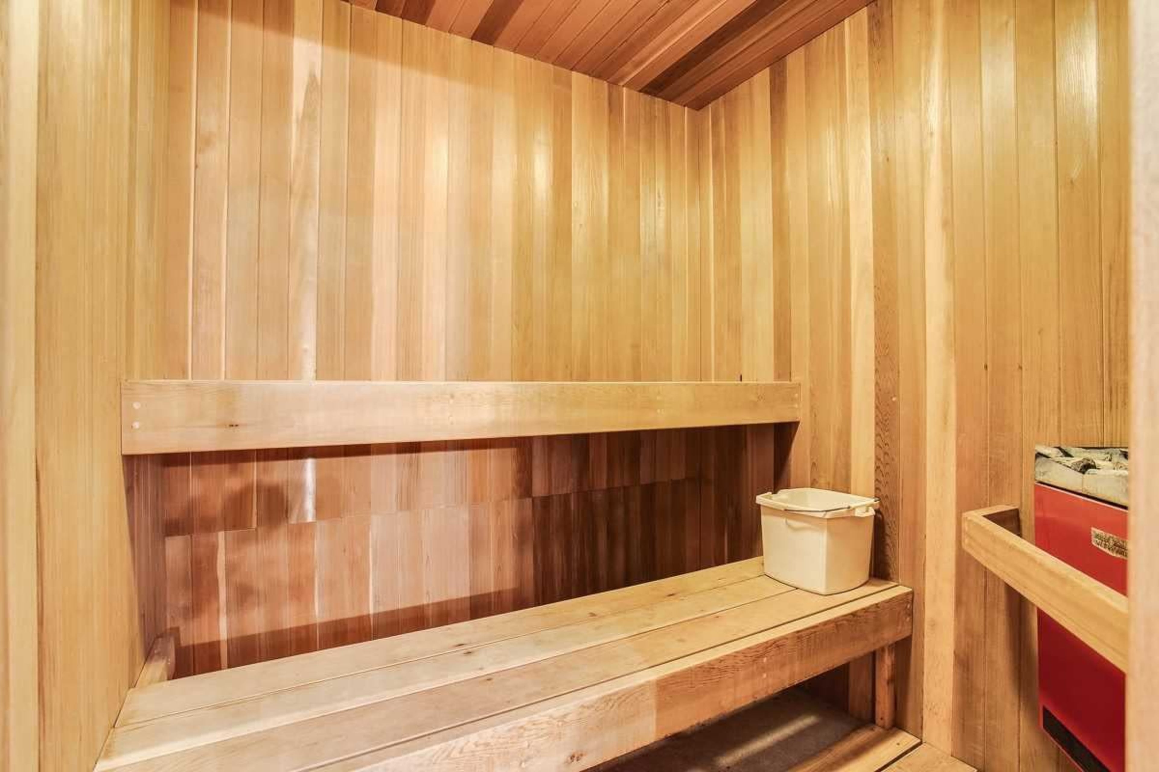 The interior of a wooden sauna with wooden bench seating and a white bucket in the corner.