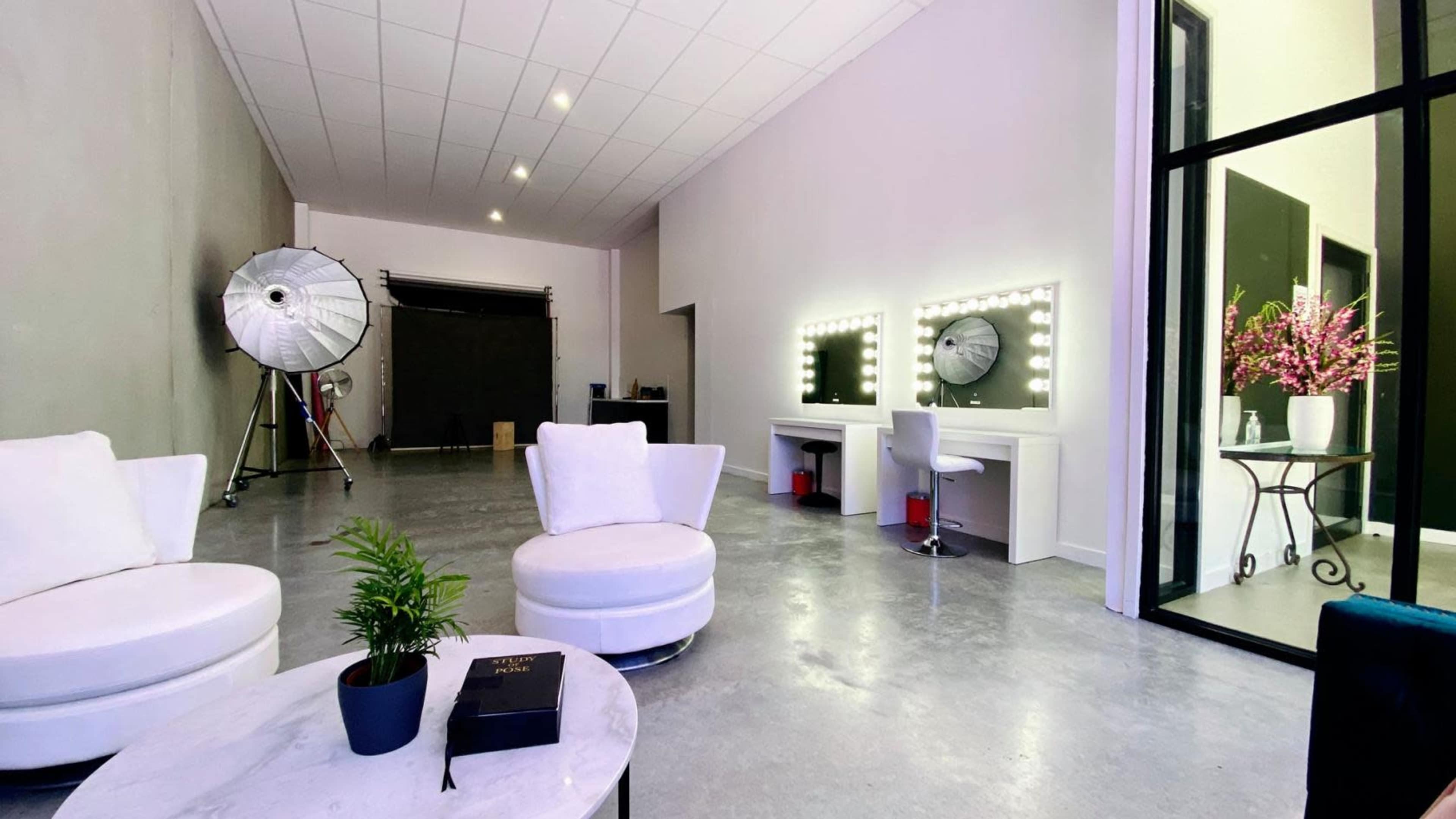 A modern interior of a cosmetic studio, featuring two illuminated mirrors, white circular chairs, a coffee table, and a large lighting setup in the background.
