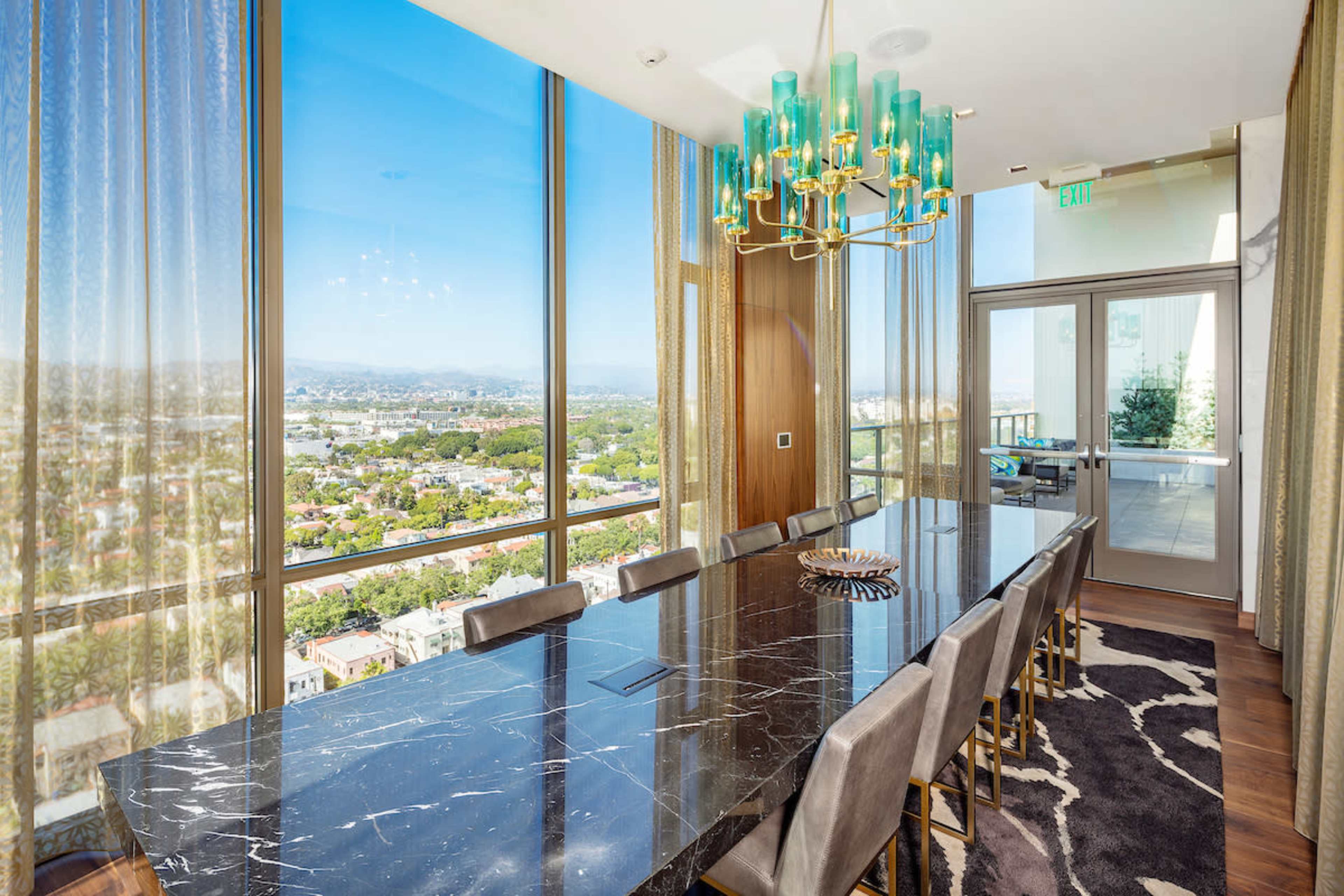 The image shows a modern dining room featuring a long black marble table surrounded by gray chairs, with large windows offering a view of the city and mountains.